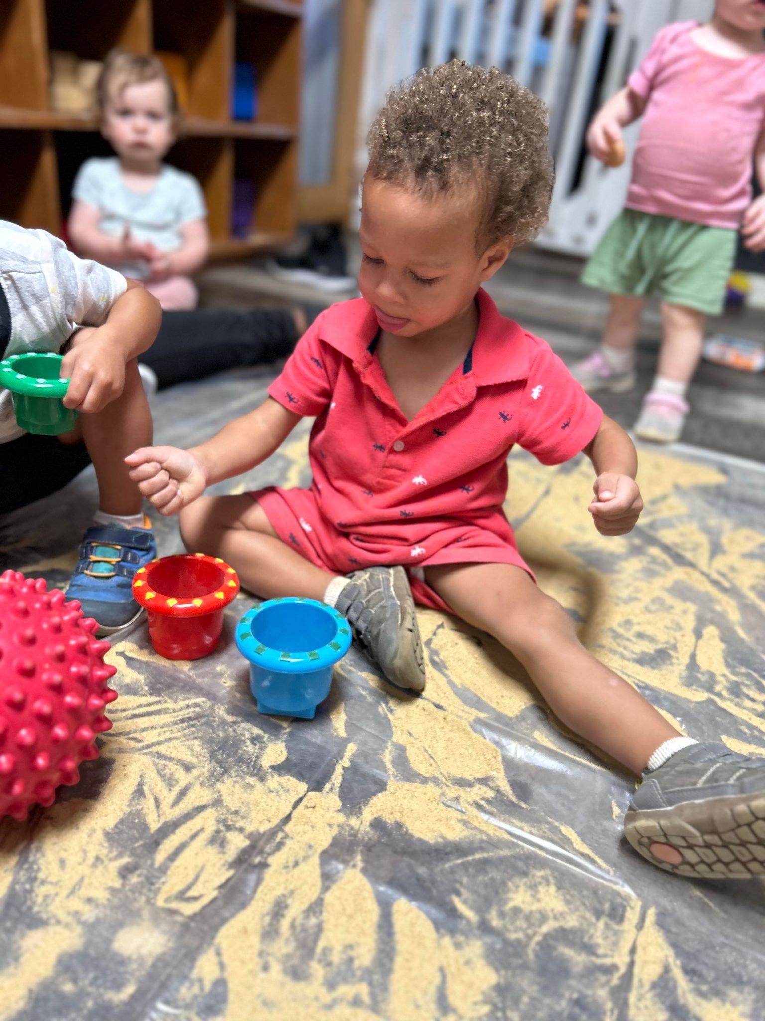 A young boy is sitting on the floor, playing with colorful toy cups and a spiky ball.
