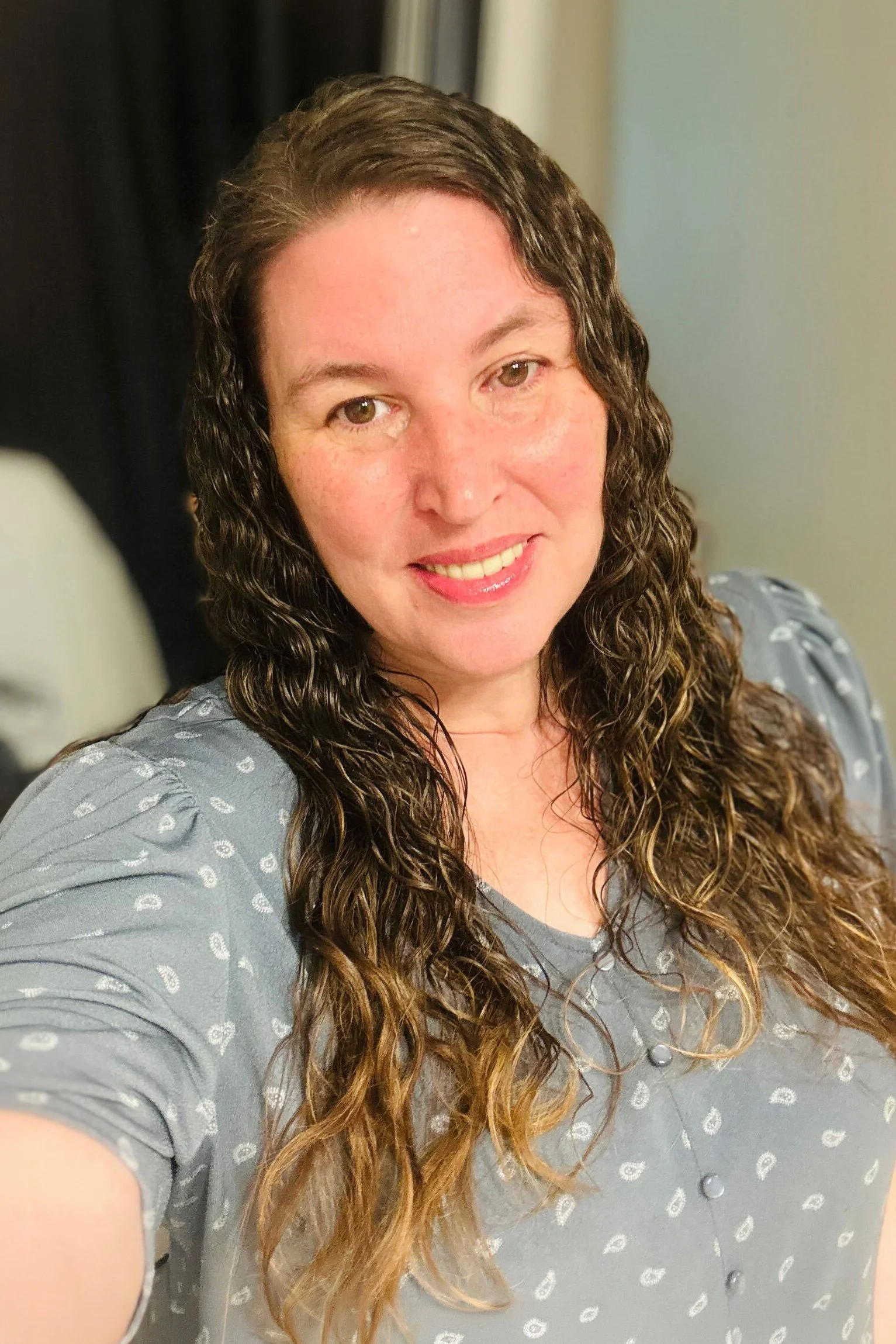 A woman with curly brown hair smiling at the camera, wearing a gray top with a pattern of small white leaves.