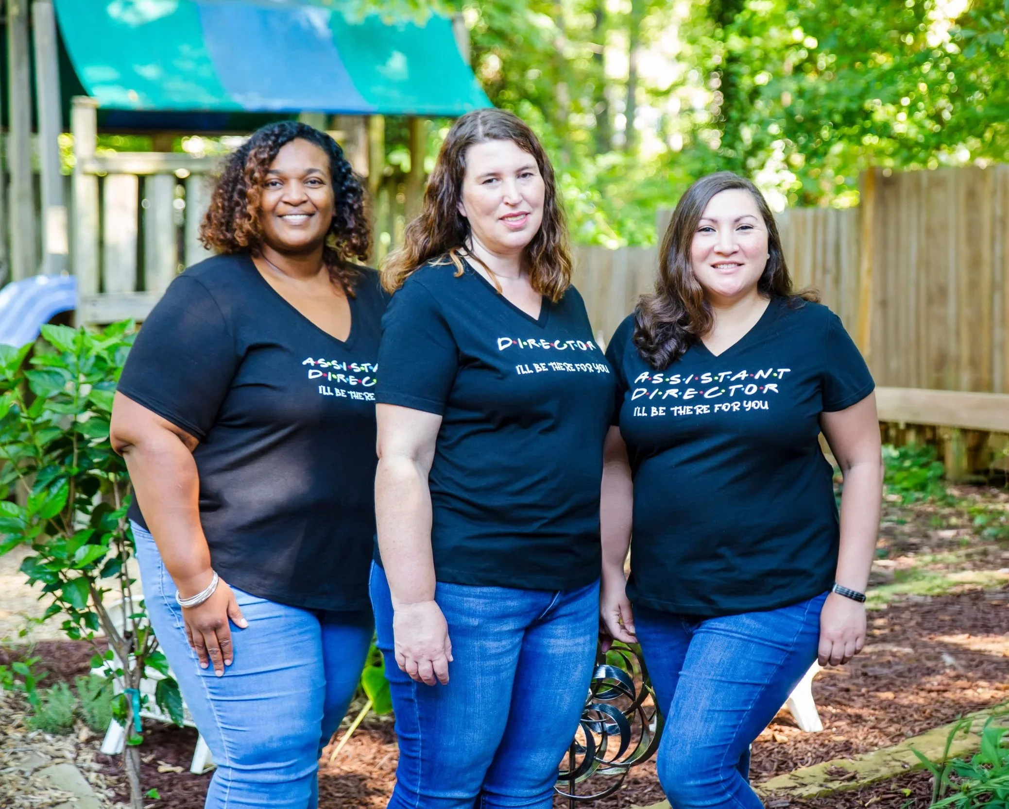 Three women standing outdoors in front of a wooden fence and green trees, all wearing black shirts with colorful text about being assistants and directors.