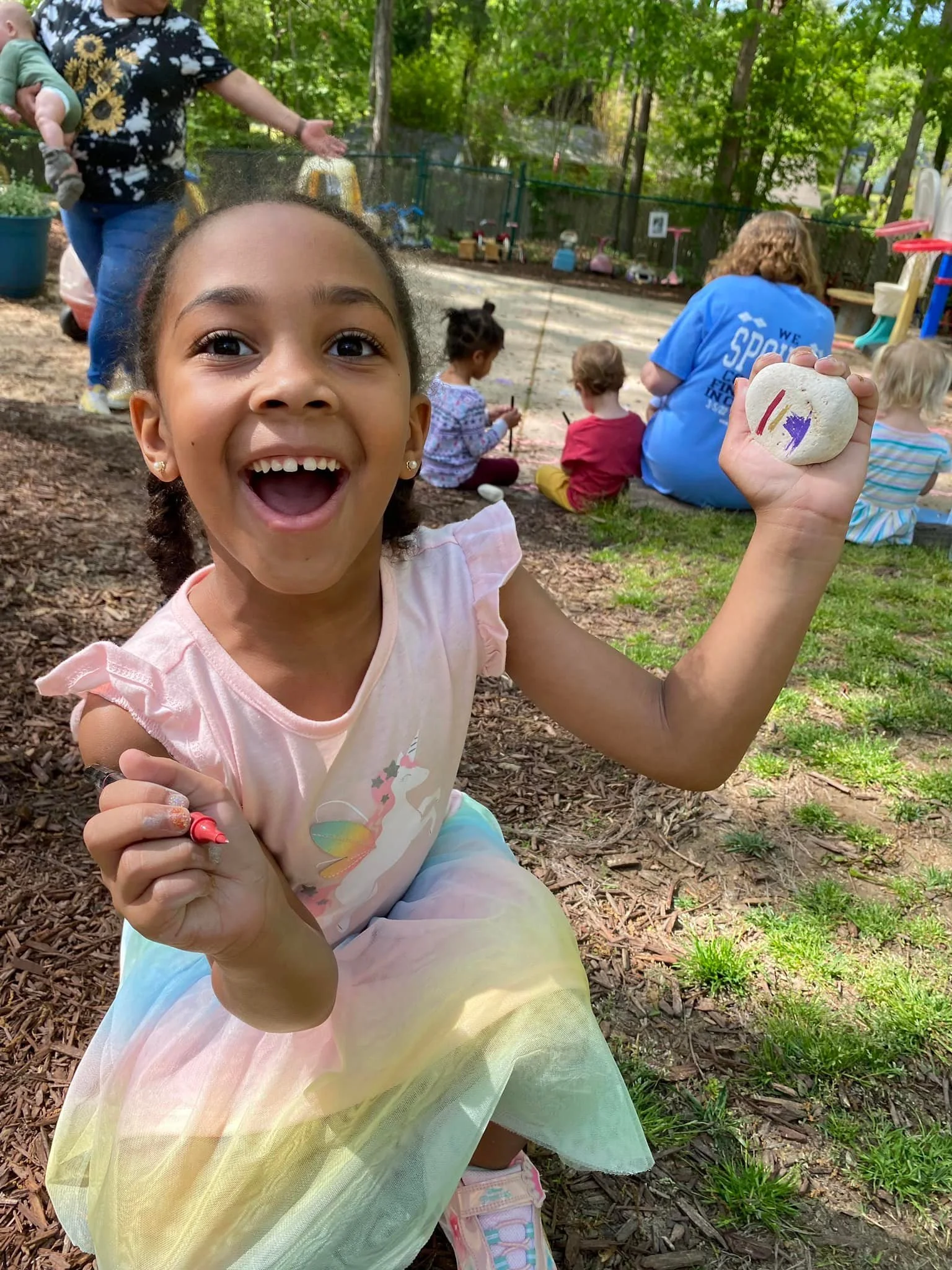 A young girl smiling happily, holding a painted rock in her right hand and a red marker in her left hand, outdoors in a park or playground with other children and an adult in the background.