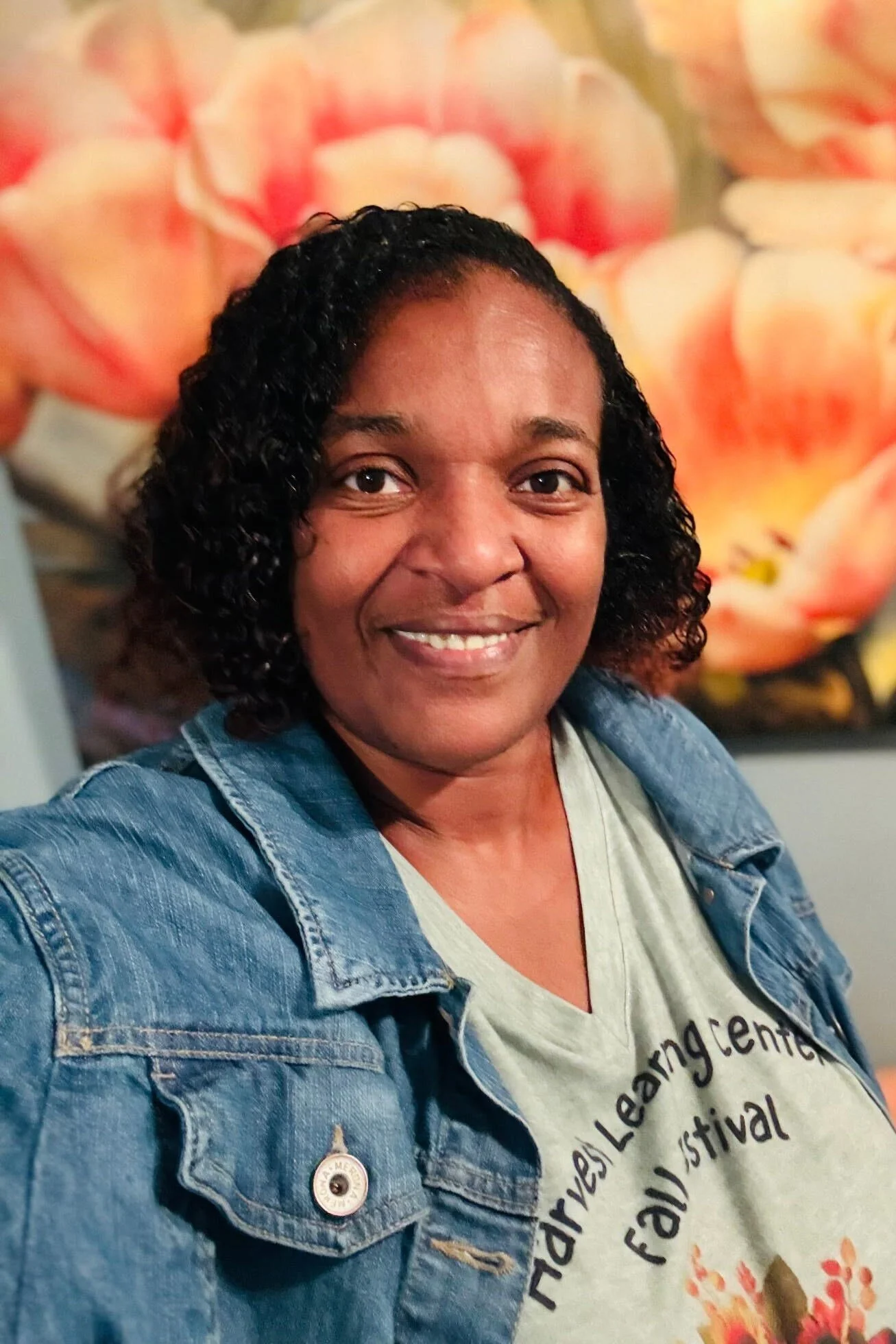 A woman with curly black hair smiling, wearing a denim jacket over a light-colored T-shirt with black text, standing in front of a floral background.