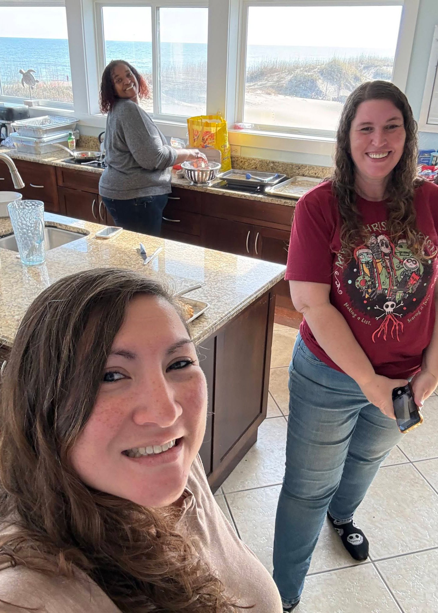 Two women smiling and taking a selfie in a kitchen with sea view outside the window. One woman has curly brown hair and freckles, and is in the foreground. The other woman has wavy brown hair, is wearing a red T-shirt with characters and graphics, and is holding a smartphone.