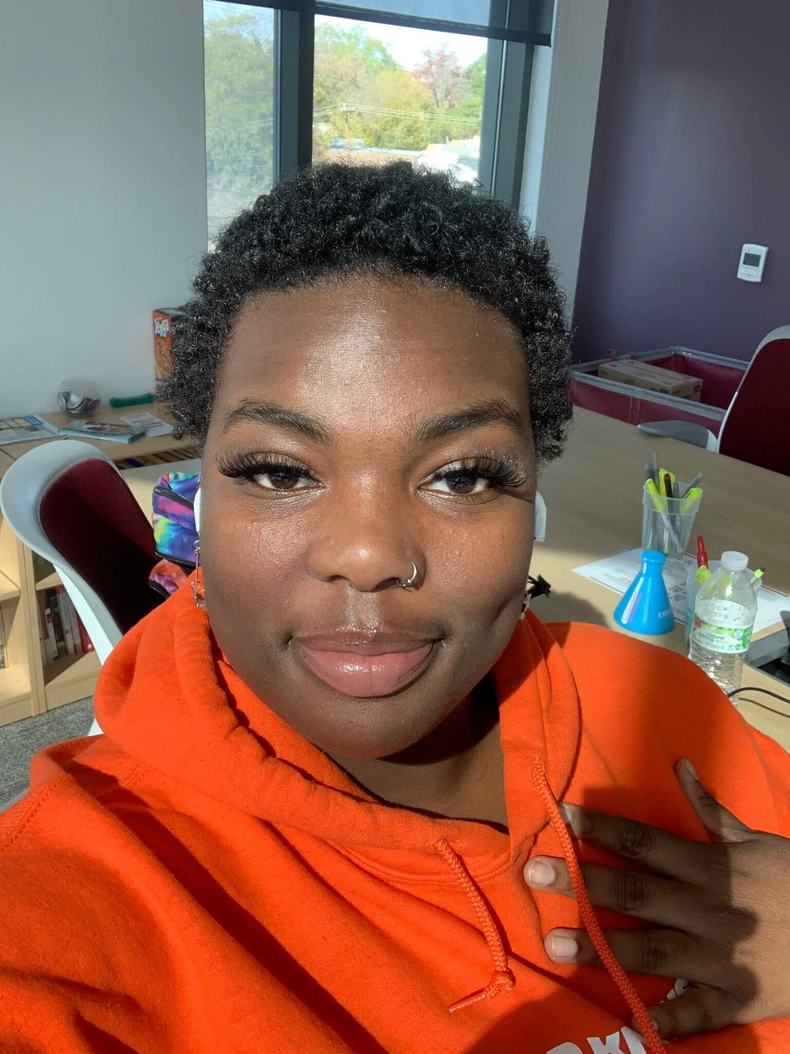 A woman with curly hair and dark skin wearing an orange hoodie, sitting in an office or classroom, smiling with her eyes closed, with a table, a chair, and laboratory equipment in the background.