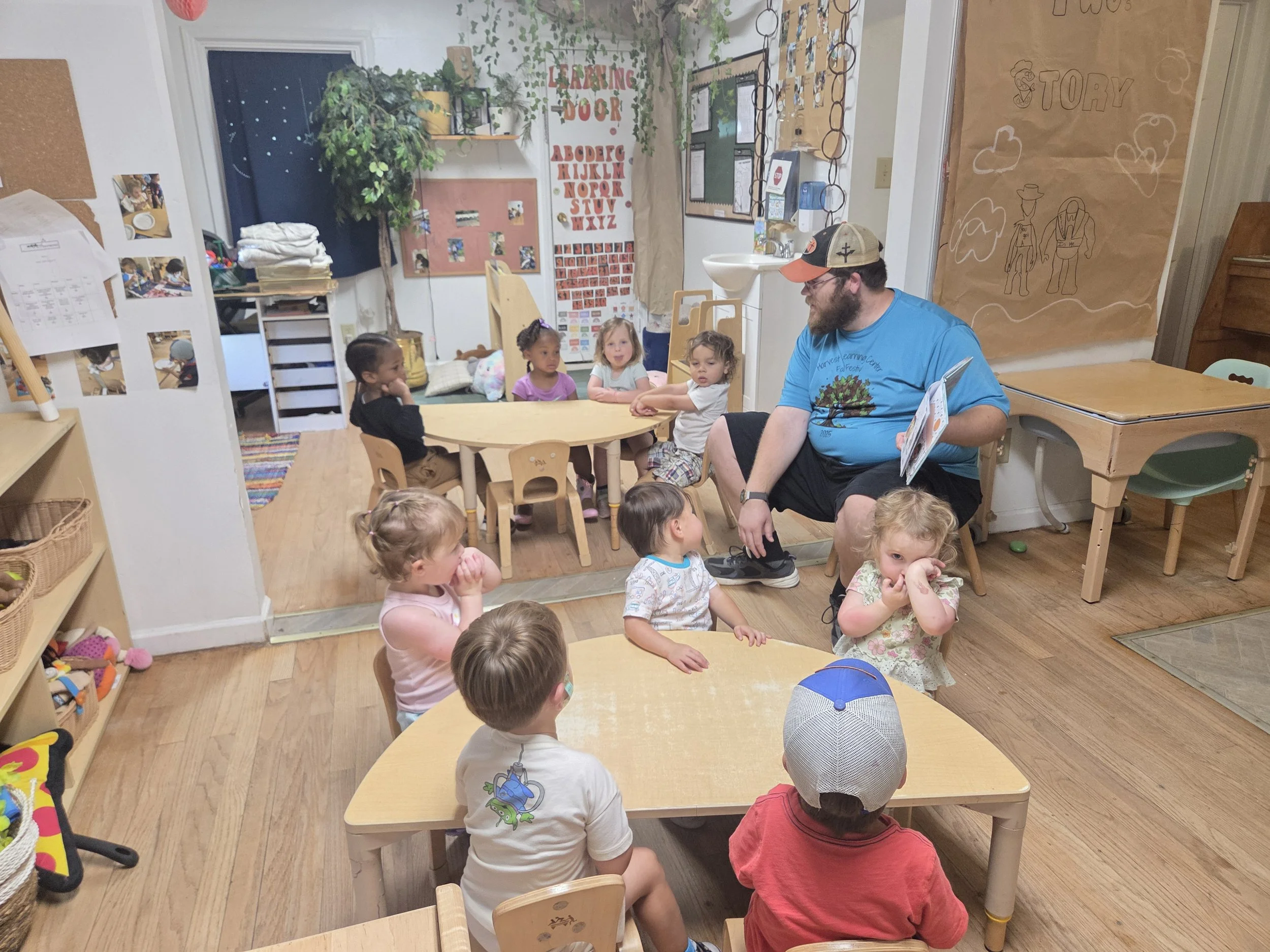 Children sitting at small tables listening to a man in a blue t-shirt and cap, in a classroom decorated with educational posters, plants, and children's artwork.