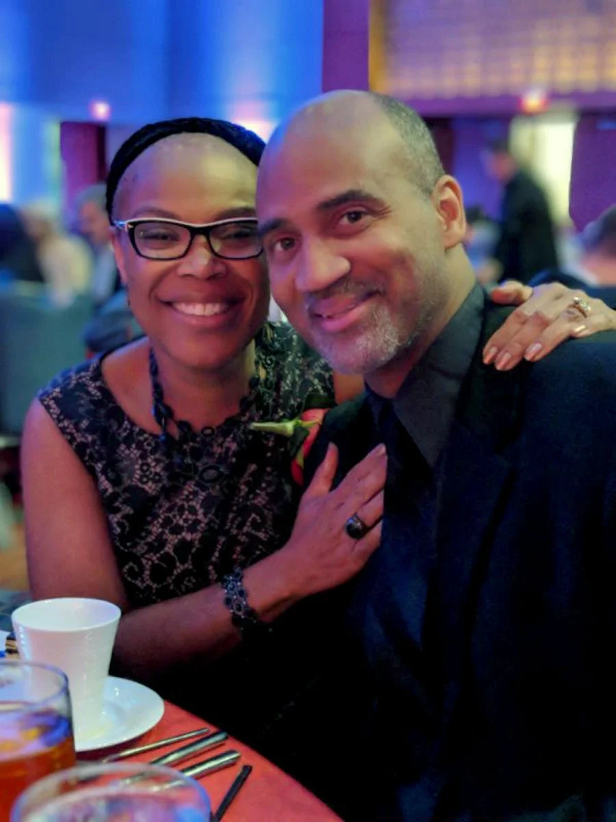 A smiling woman with glasses and a man with a shaved head and beard, sitting at a table at a social event or celebration, with blue and purple lighting in the background.
