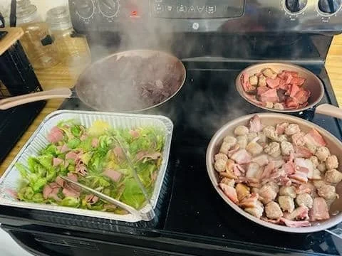 Stove with multiple pans and dishes being cooked, including a large pot, a skillet with chopped meat, and a rectangular dish with lettuce, ham, and sliced vegetables.