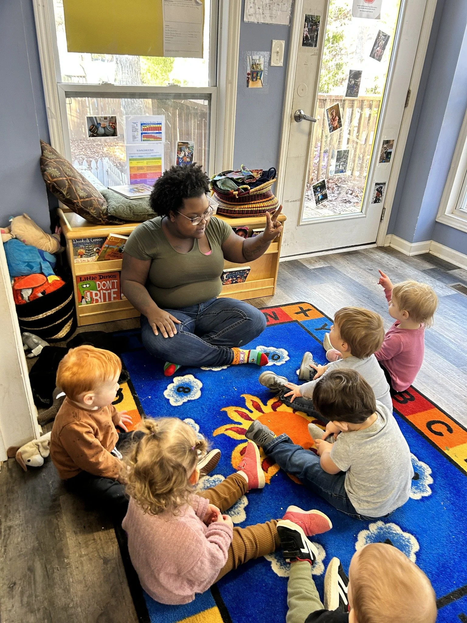 A woman sitting on a colorful rug in a classroom with six young children, engaging in a story or lesson. The room has a wooden shelf with books and bags, large windows, and a door with photos and drawings attached.