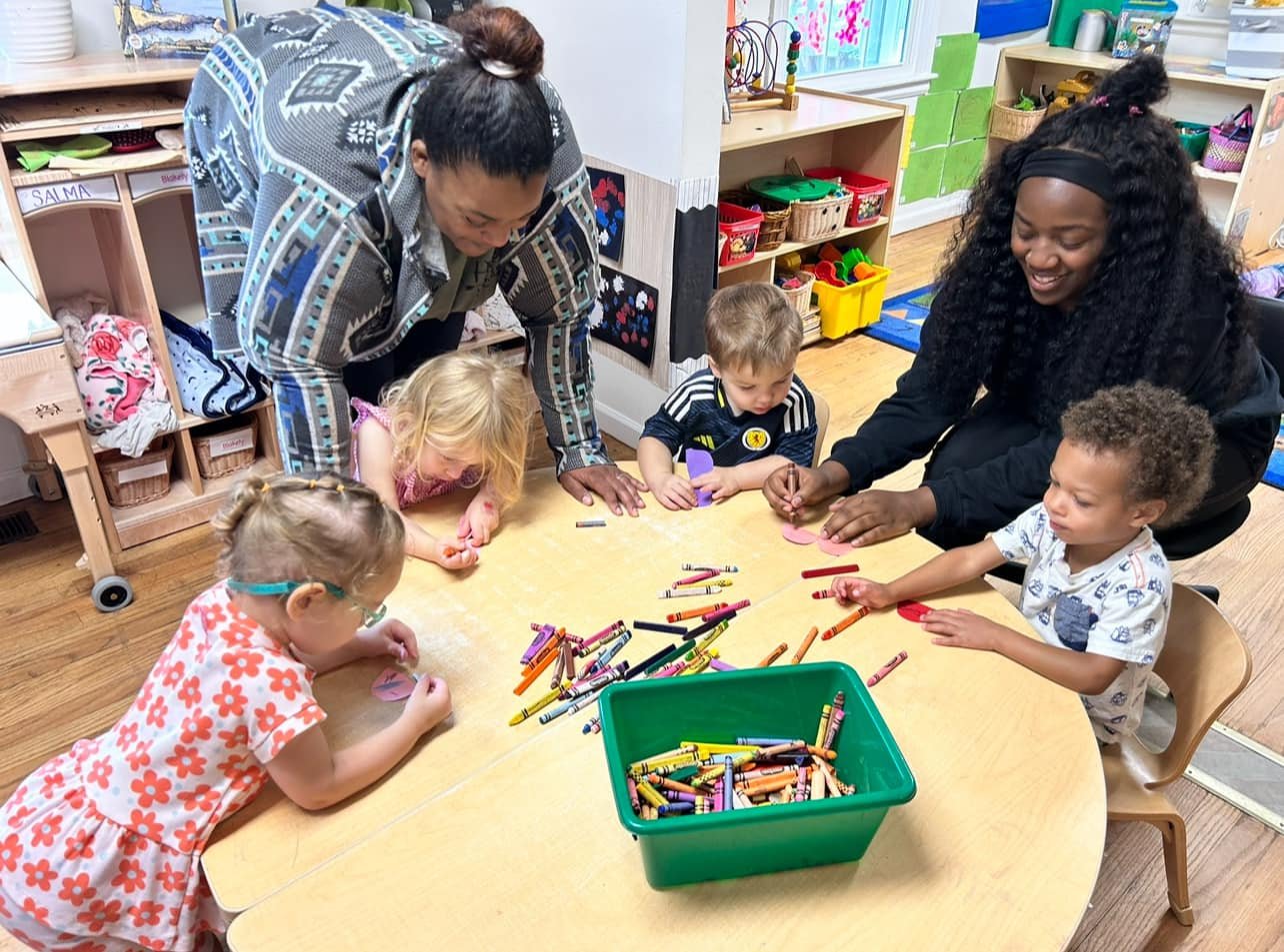 Two teachers and four young children sitting around a wooden table in a classroom, engaged in a coloring activity with crayons and paper.