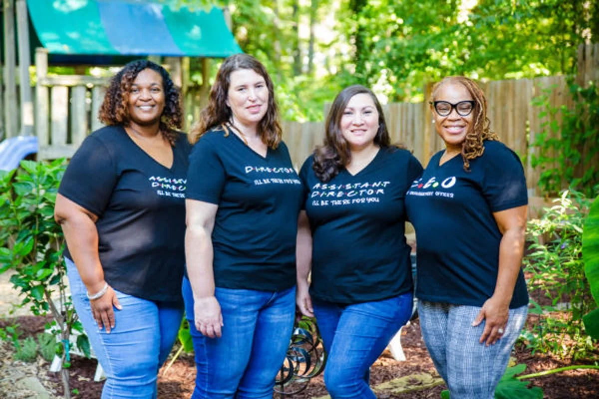 Mrs. Lashanda, Mrs. Ginger, Mrs. Ashley, and Mrs. Lynette standing outdoors in front of a wooden fence and lush green trees, wearing matching HLC shirts.