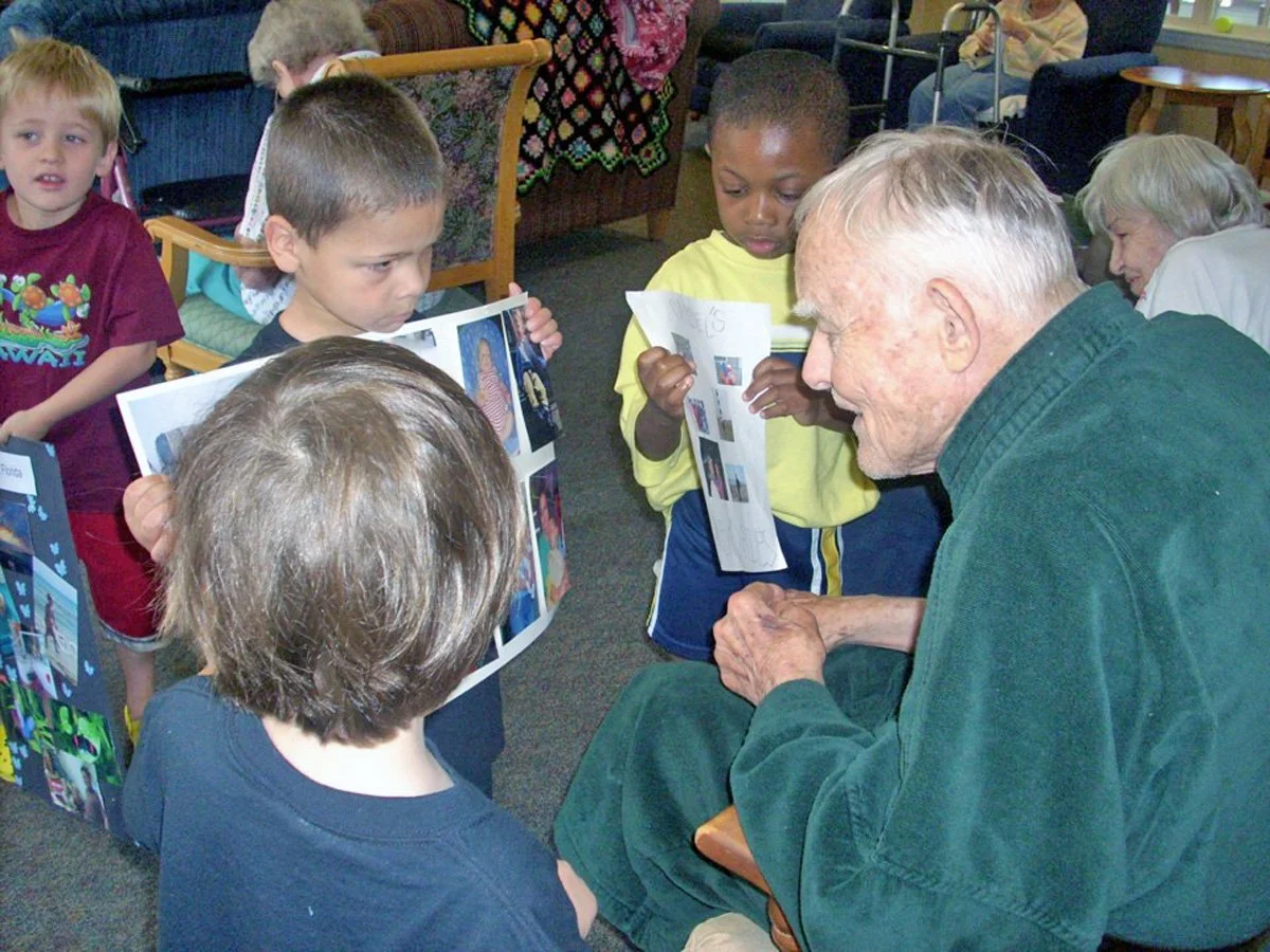A group of young children and an elderly man gathered together, looking at a photo album or pictures. The kids are holding open books or picture sheets, and they appear engaged with the elderly man who has white hair and is smiling.