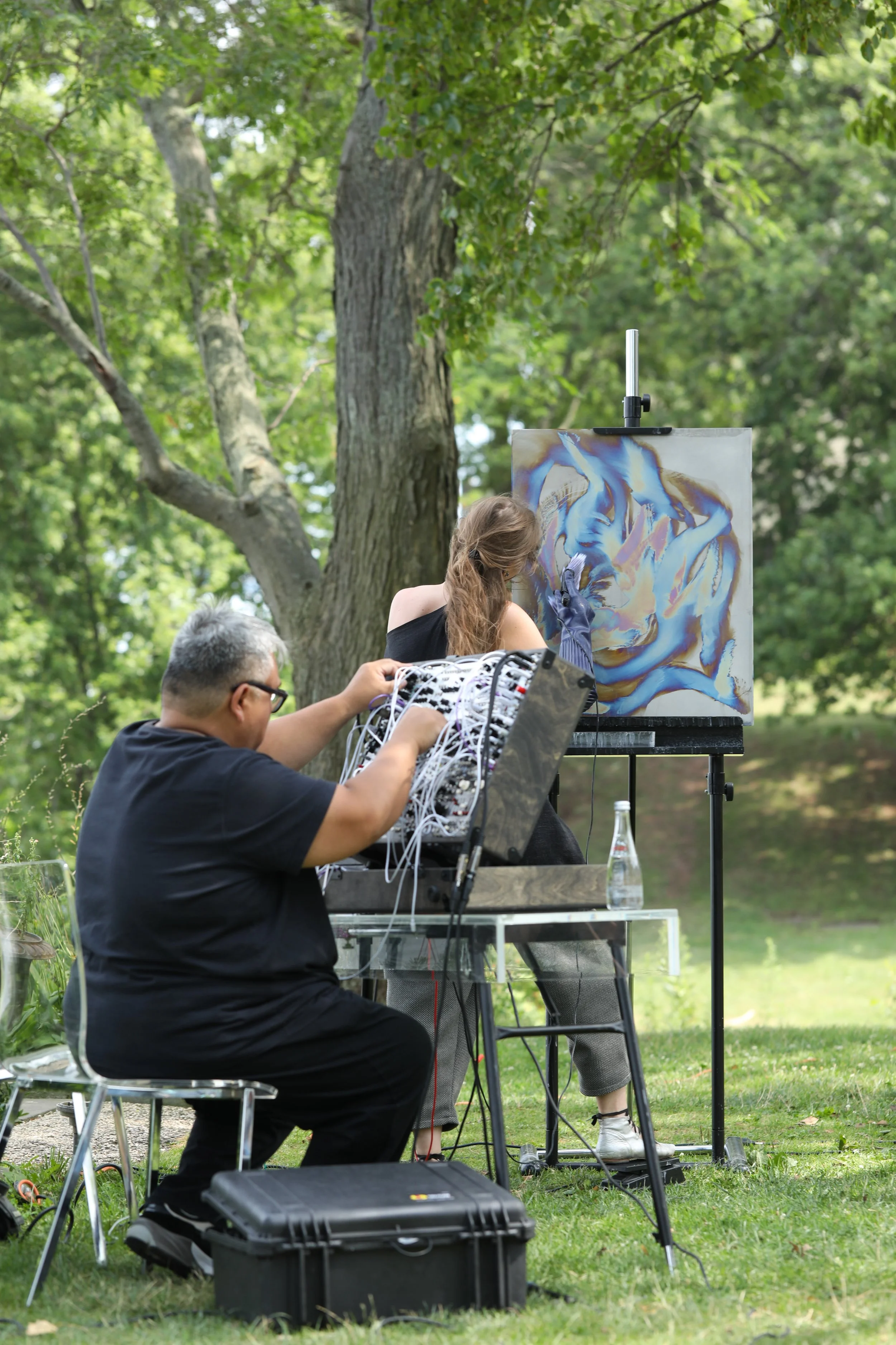 A woman is painting an abstract artwork on a canvas outdoors in a park, with a man managing electronic equipment nearby.
