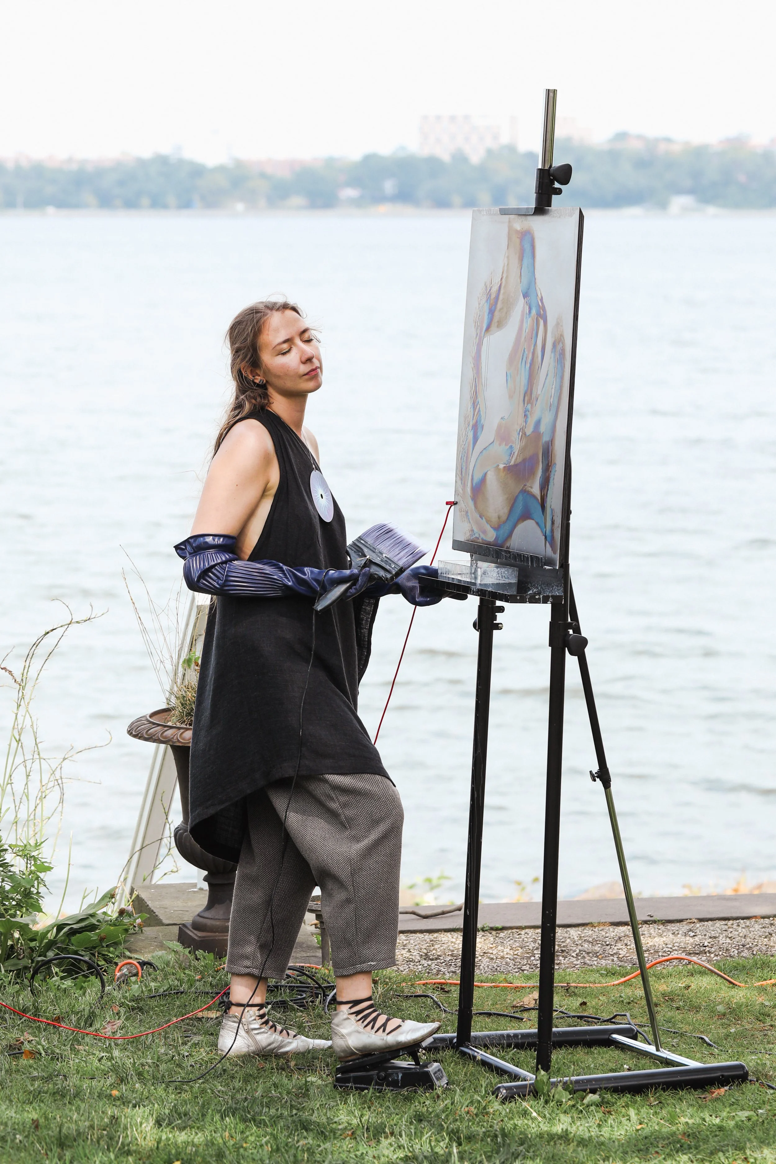A woman painting outdoors by a body of water, standing on grass, with painting supplies and an easel.