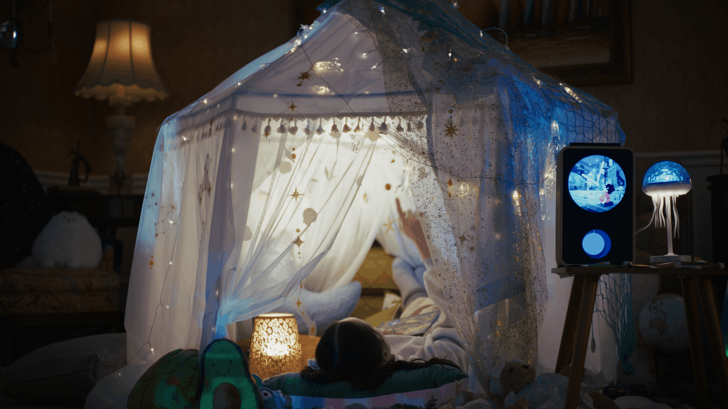 Child lying in a blanket tent decorated with stars and fairy lights, watching a tablet. Nearby, a bedside lamp and a night light shaped like a jellyfish are on a side table, creating a cozy, dreamy atmosphere.
