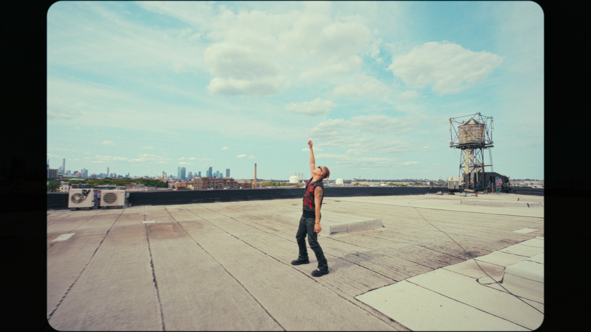 A person standing on a city rooftop under a partly cloudy sky, holding their head back and pointing upward with their right arm. They are wearing a sleeveless shirt, jeans, and sunglasses, with visible tattoos on their arms. The rooftop has a flat surface with rooftop equipment and a city skyline in the background.