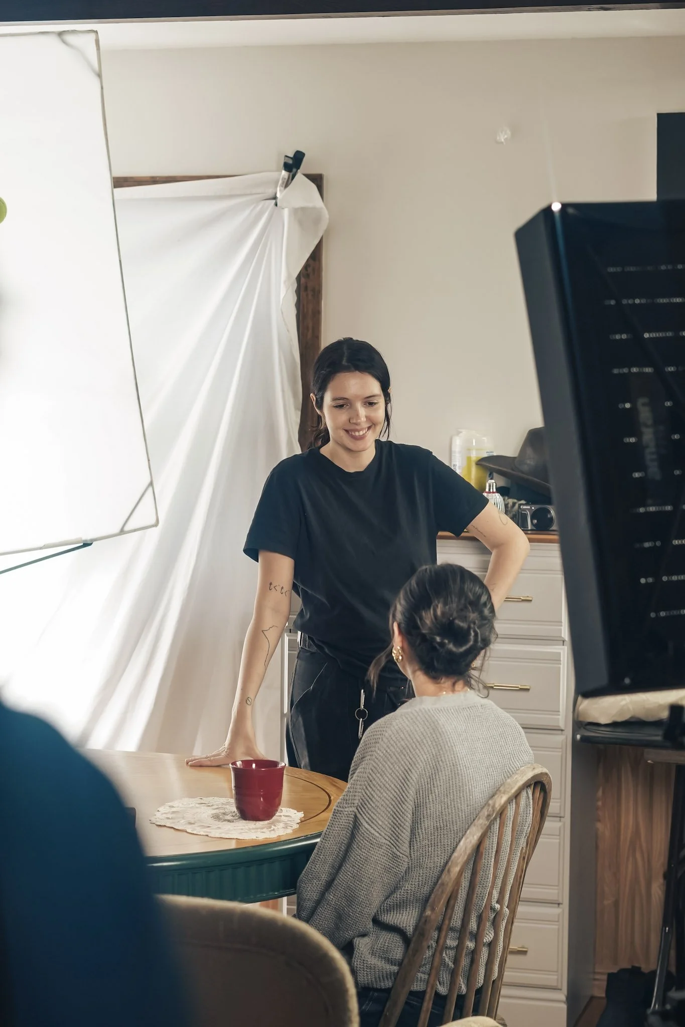 A woman in a black t-shirt is standing and smiling at another woman with dark hair in a bun, who is sitting at a wooden dining table. The sitting woman is wearing a gray sweater and gold earrings. There is a red mug on a doily on the table. The background shows white drawers, a camera, and some appliances.
