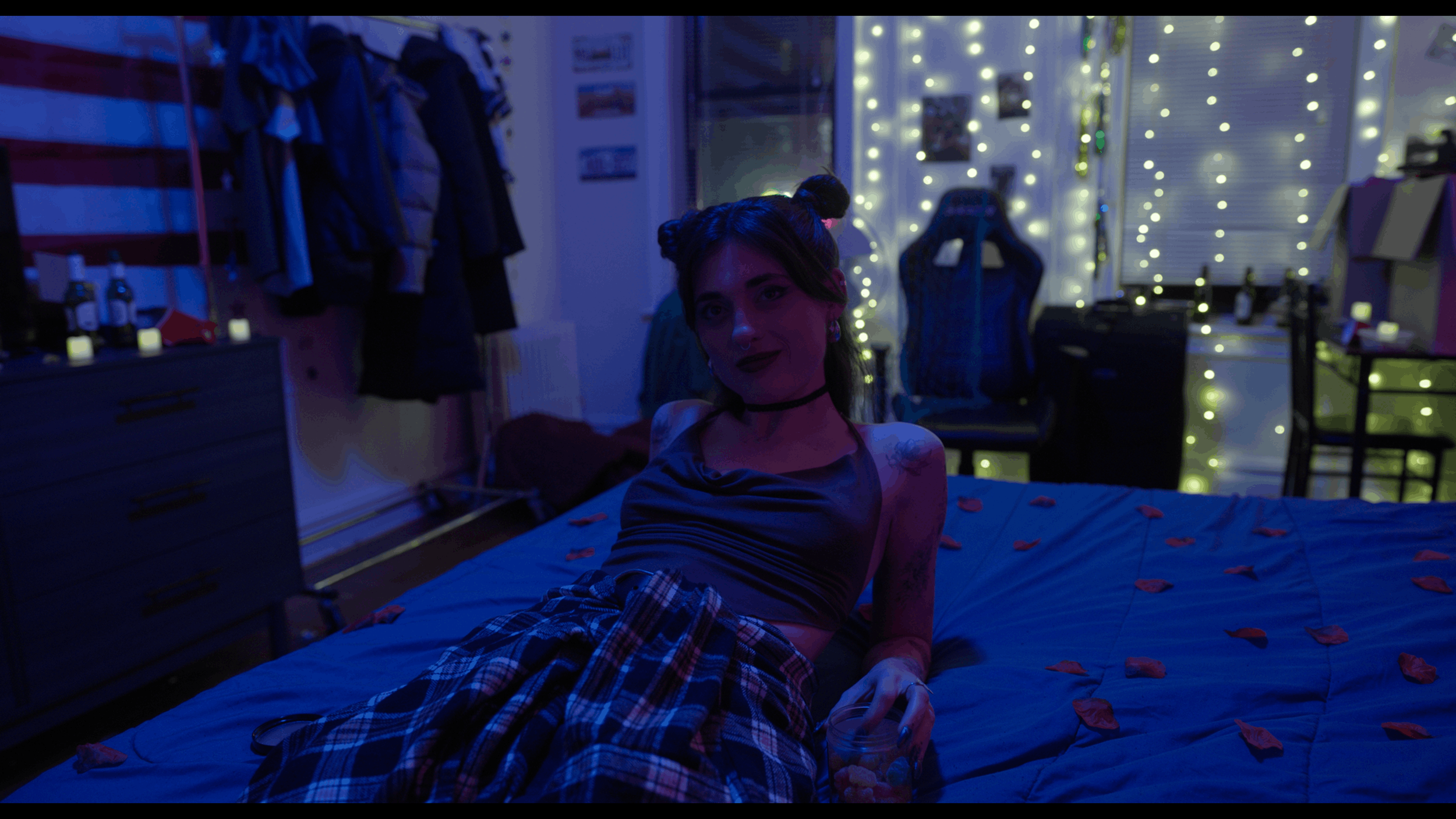 A woman with dark hair styled in buns, wearing a choker and dress, lying on a bed in a colorful, dimly lit room decorated with fairy lights, with a glass of snacks in her hand, and clothes hanging in the background.