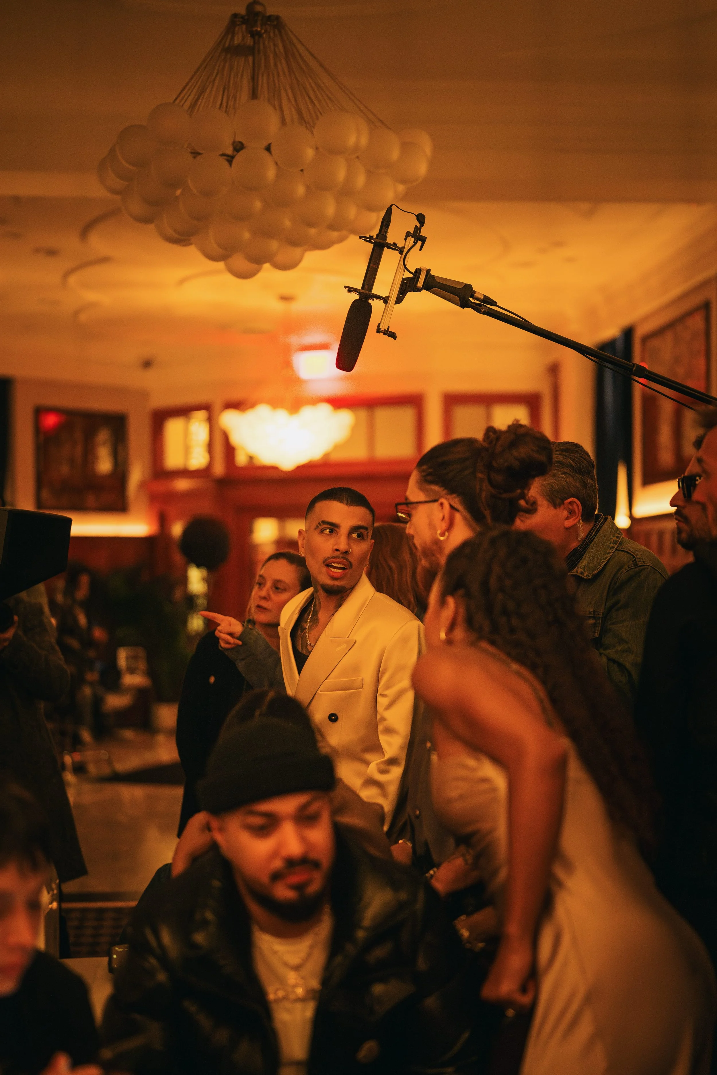 People gathered in a warmly lit indoor space with a crew member and microphone overhead, likely on a film or music video set.