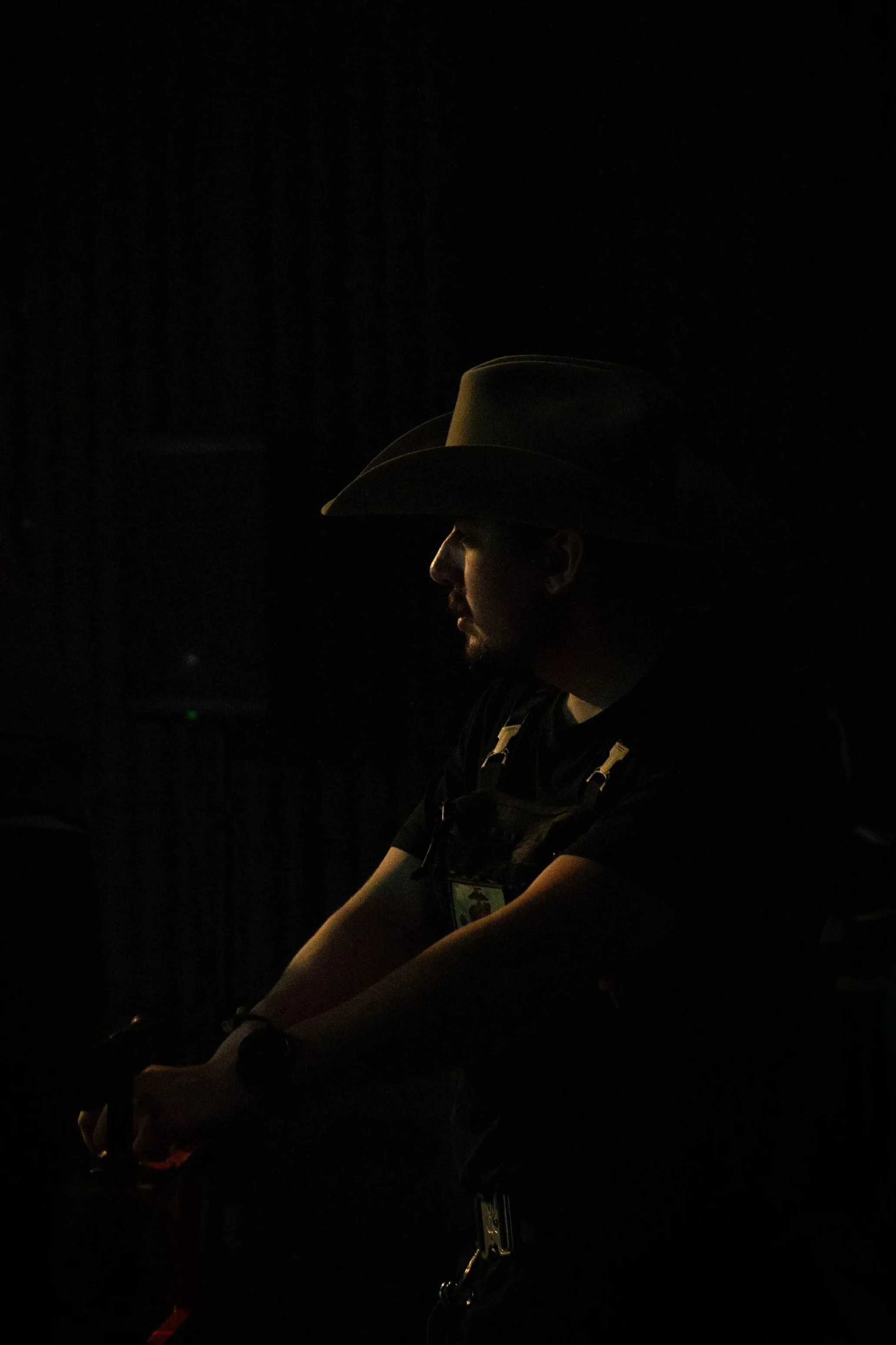 A person wearing a cowboy hat is sitting in a dark environment, with minimal lighting highlighting their profile.