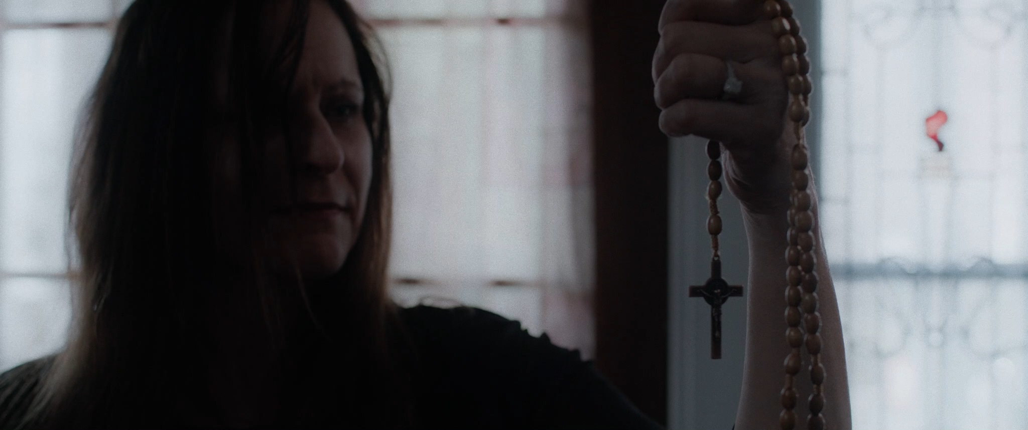 A woman with long hair is holding up a string of wooden rosary beads with a cross, in a dimly lit room with light coming through a window in the background.