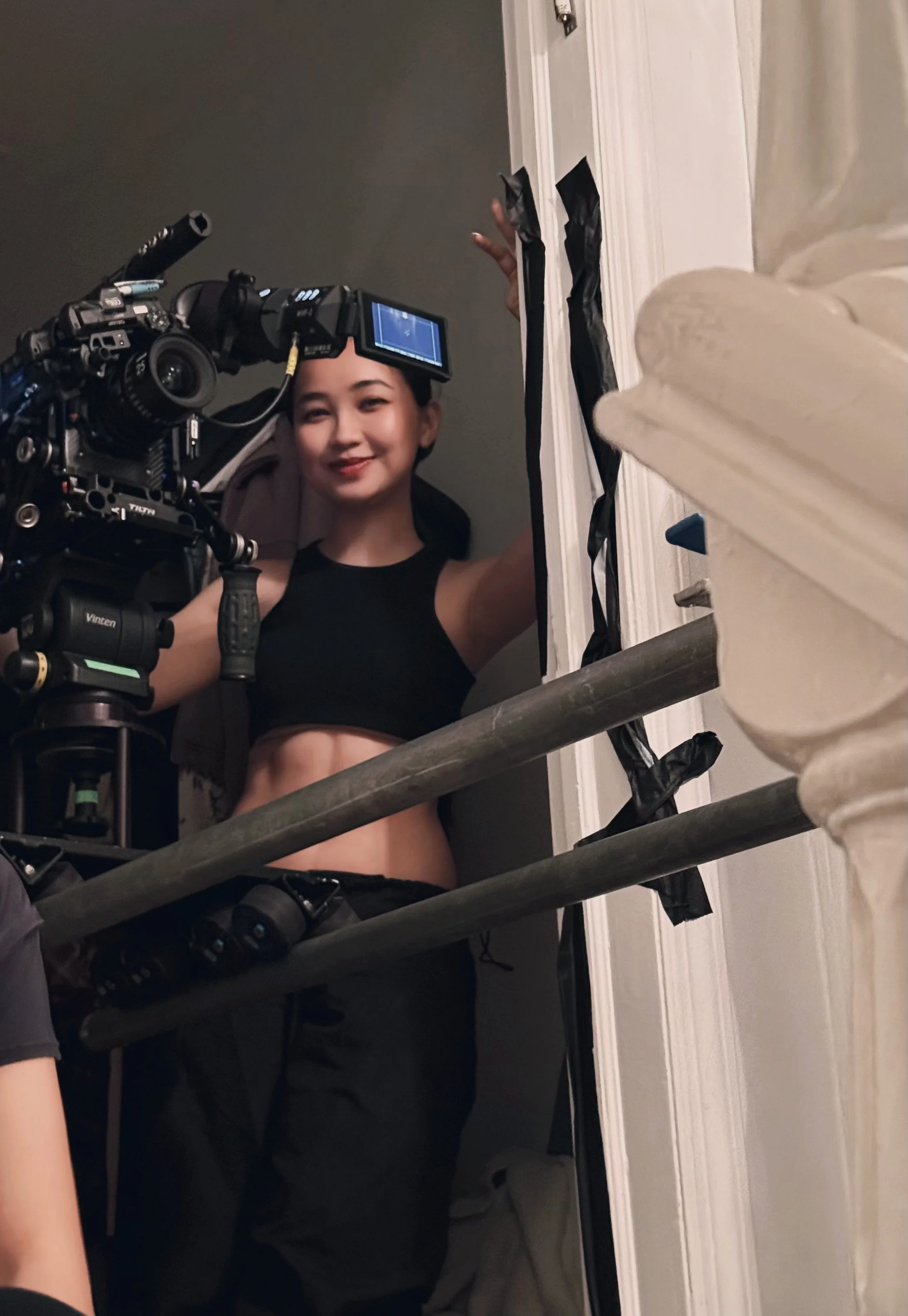 A young woman dressed in a black sports bra during a film or photo shoot with a professional camera, smiling and looking at the camera, in a small enclosed space.