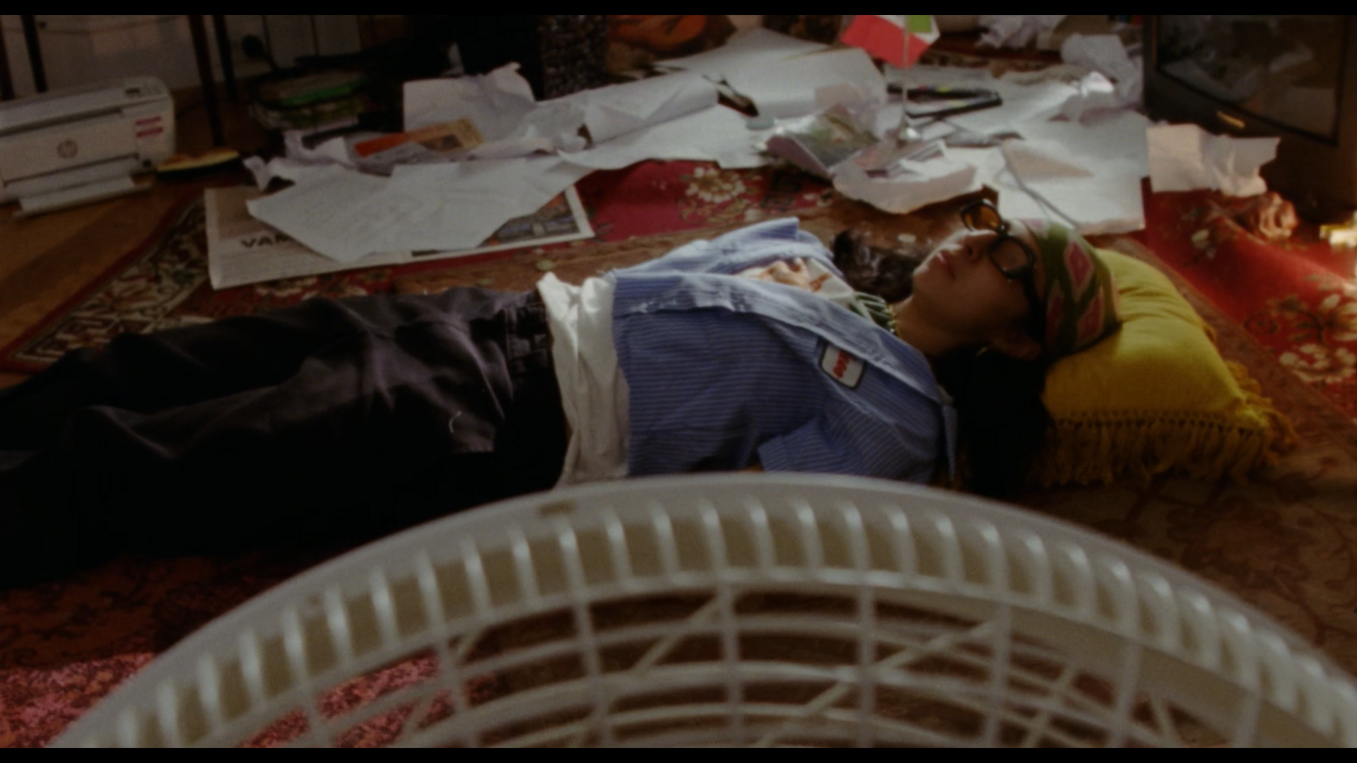 A person lying on their back on the floor, surrounded by scattered papers and cluttered items, with a blanket over part of their body, resting on a patterned carpet.