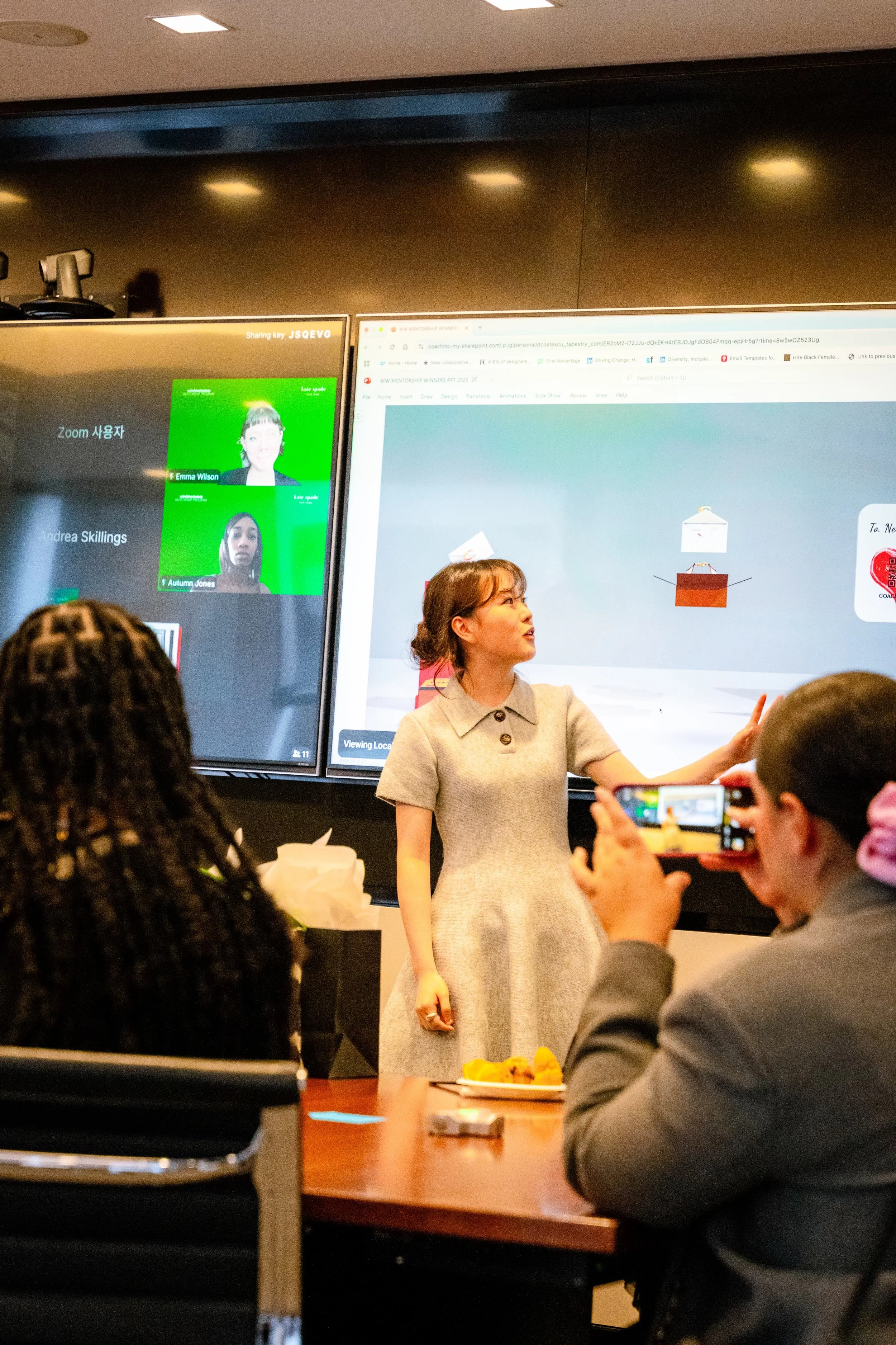 A woman in a beige dress giving a presentation in front of a large screen, with several people seated and recording the presentation on their phones.