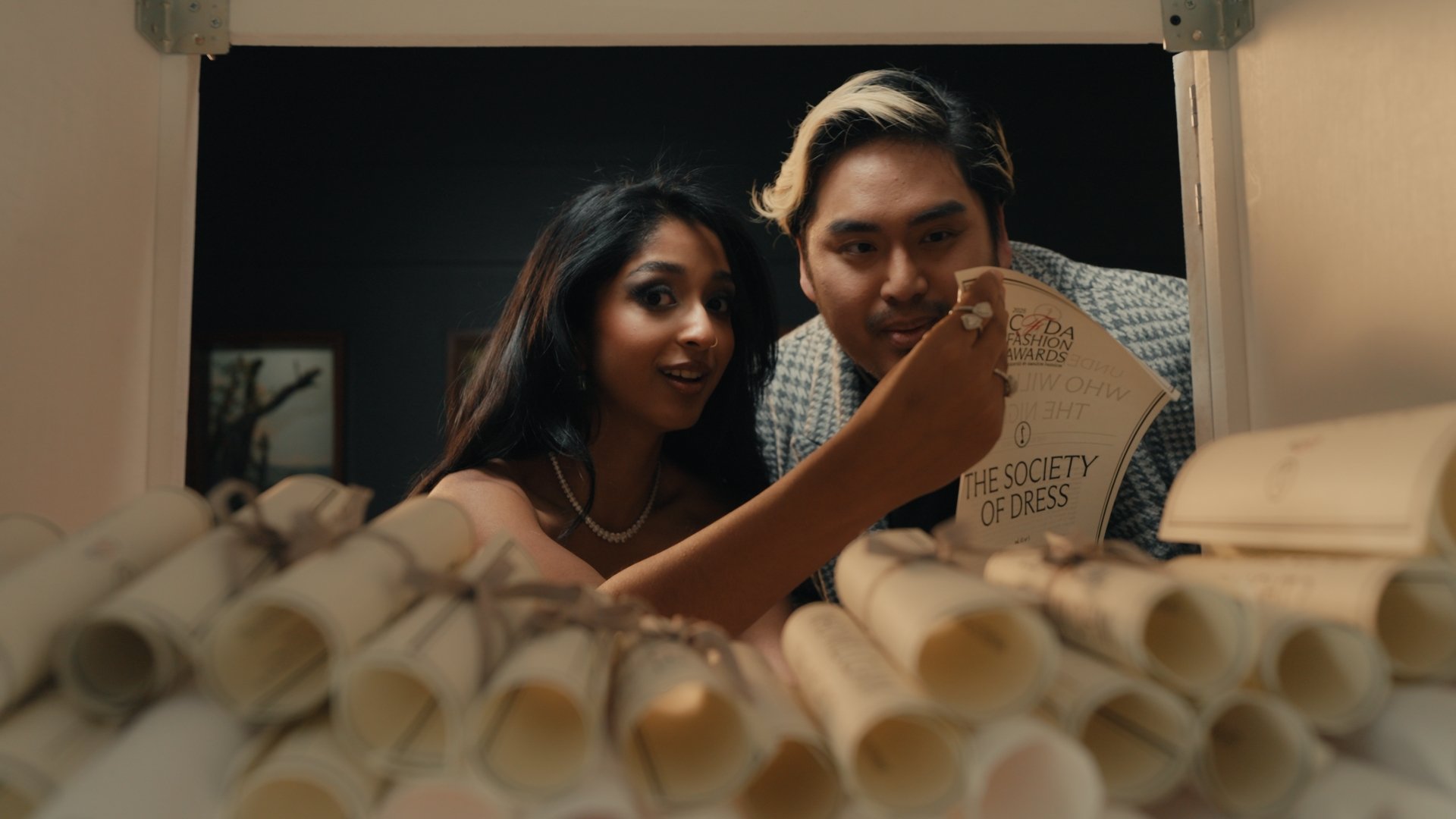 A man and woman looking into a box filled with rolled-up papers or scrolls, with the woman holding a paper that says 'THE SOCIETY OF DRESS'.