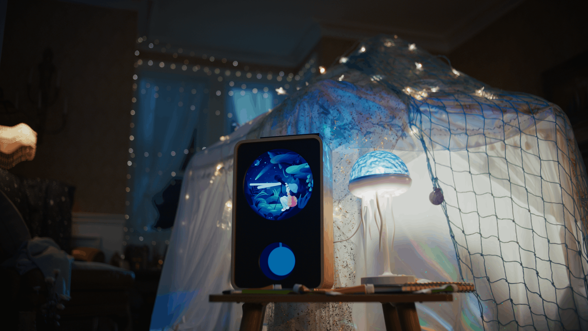 A night scene featuring a starry sky with fairylights, a chair with a net canopy decorated with fairy lights, a small table with a lamp resembling a jellyfish, and a tablet showing a scene of a girl fishing with whales. The room has blue and white tones with cozy decor.