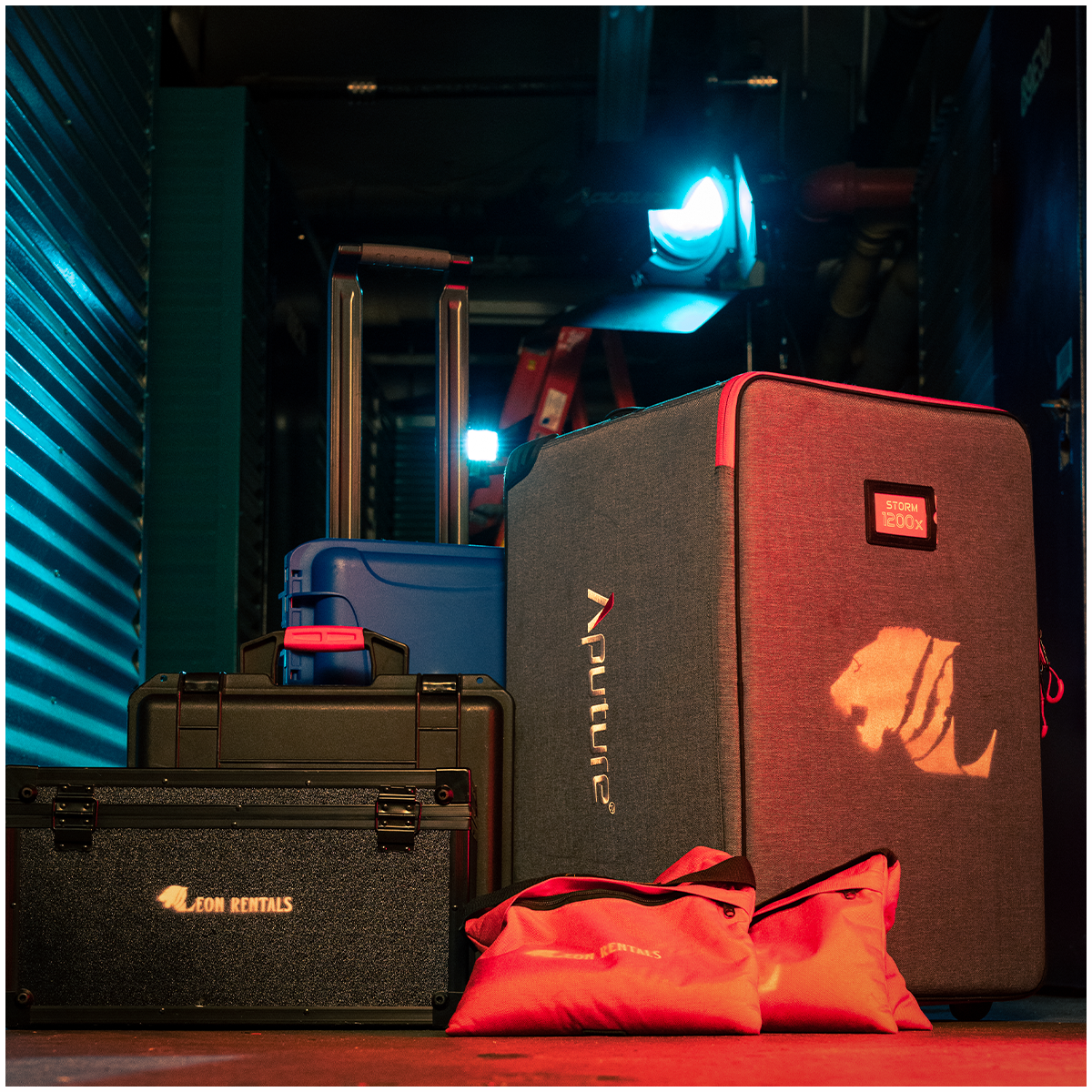 Luggage and equipment stacked in a dimly lit storage unit, including a large grey case with lion logo, a blue suitcase, a black case with illuminated 'Aston Rentals' logo, and red bags, illuminated by blue and orange lighting.