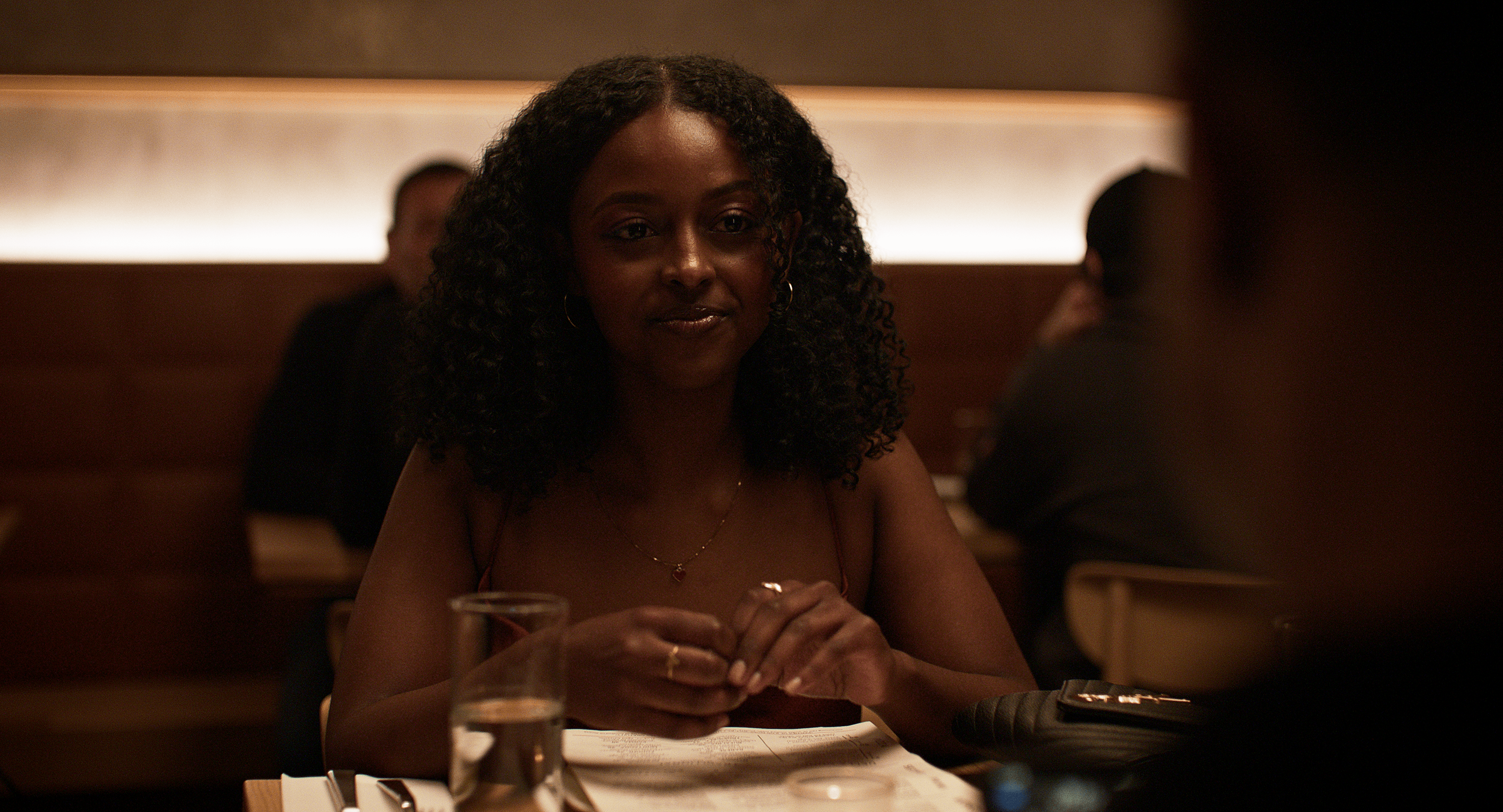 A woman with curly hair, wearing a spaghetti strap top, sitting at a restaurant table with a glass of water and a menu, looking at someone in front of her.