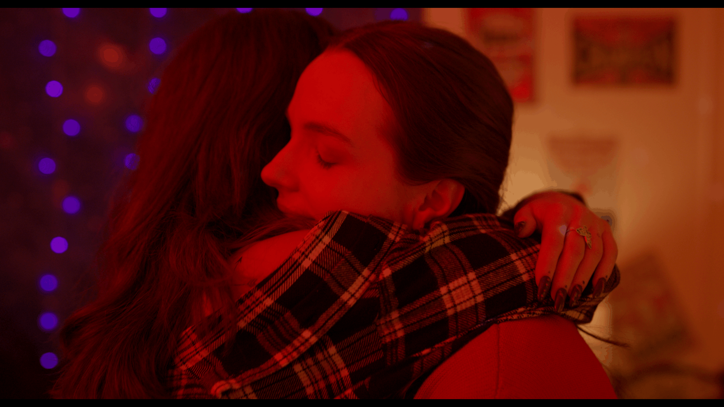 Two women hugging tightly in a room decorated with Christmas lights, one with long curly hair and the other with dark straight hair, both with closed eyes, in a warm, intimate moment.