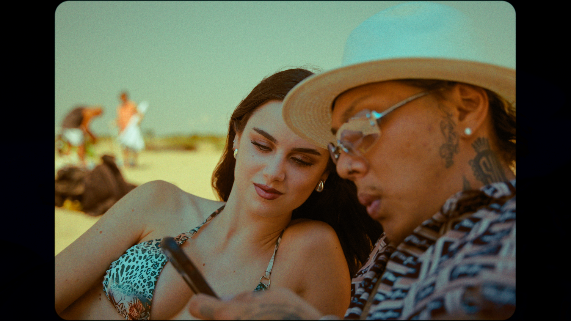 A woman with dark hair and a bikini top looking at a phone held by a man with a hat and tattoos, on a beach with others in the background.