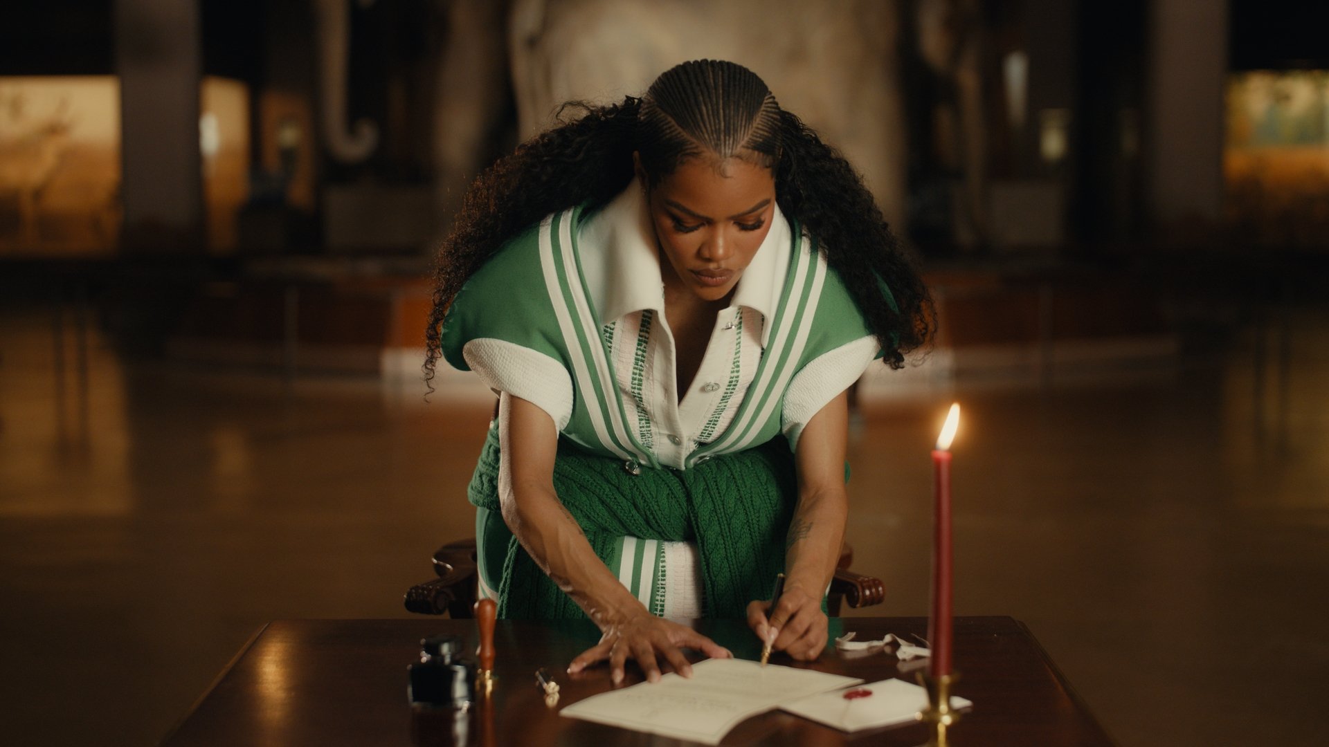 A woman with long, curly hair in a retro hairstyle, wearing a vintage green and white varsity jacket, is leaning over a desk in a dimly lit room, writing in a large open book. There are ink and writing utensils on the desk, and a lit red candle.