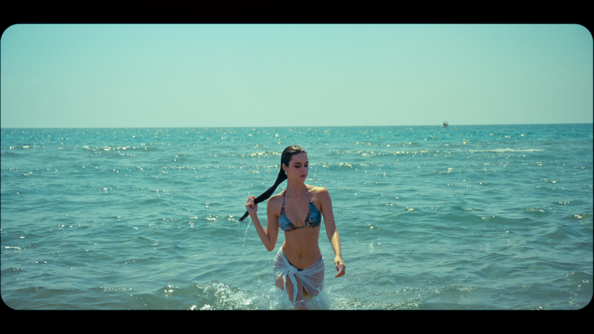 A woman in a leopard-print bikini and semi-sheer white skirt walking out of the ocean with long dark wet hair, in a sunny beach scene.