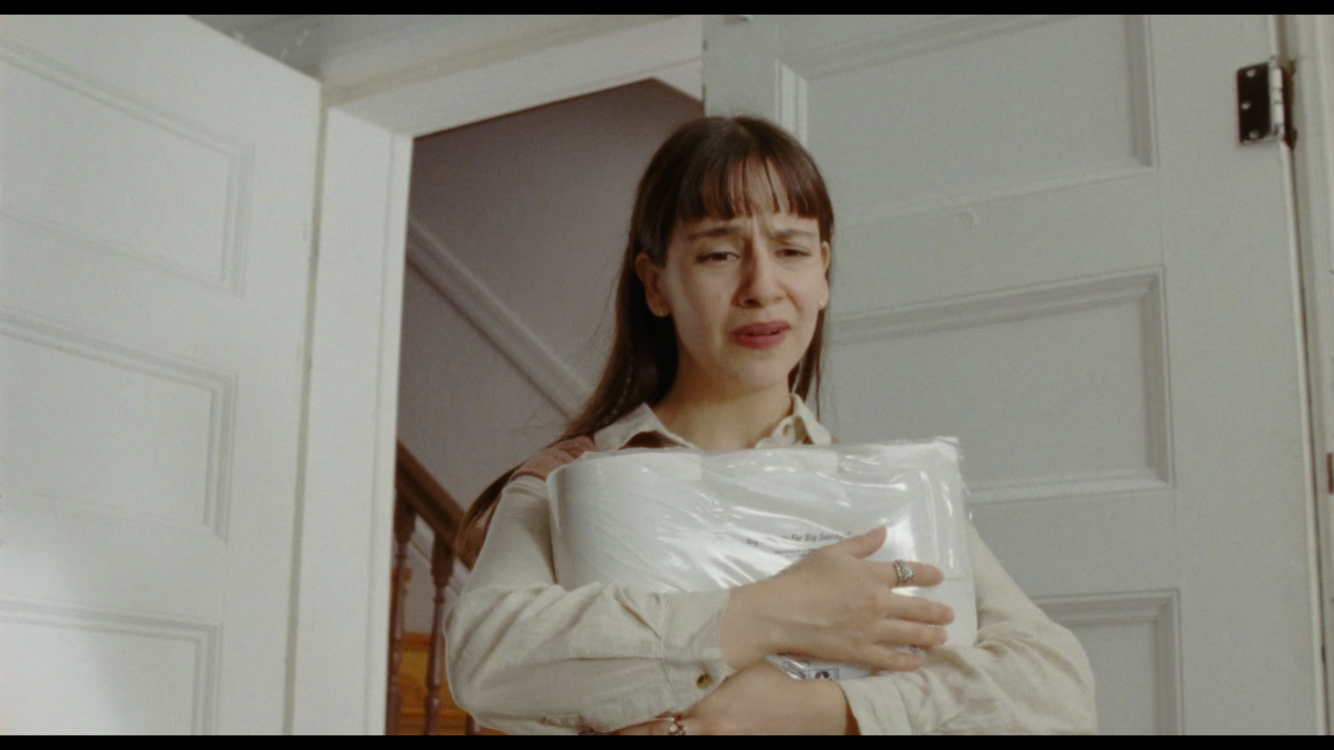 A woman with brown hair and bangs standing in a doorway, holding a large roll of toilet paper wrapped in plastic, with a distressed facial expression.
