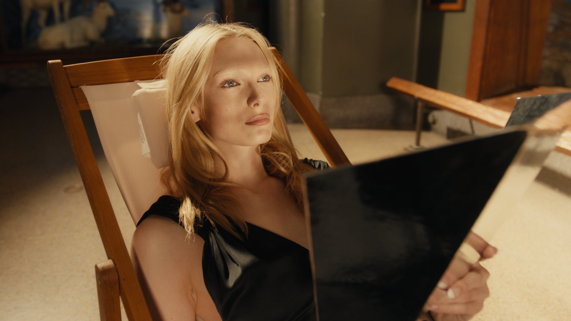 A woman with long red hair and a black top is reclining in a wooden lounge chair, holding an electronic device, possibly a tablet, with a focused expression.