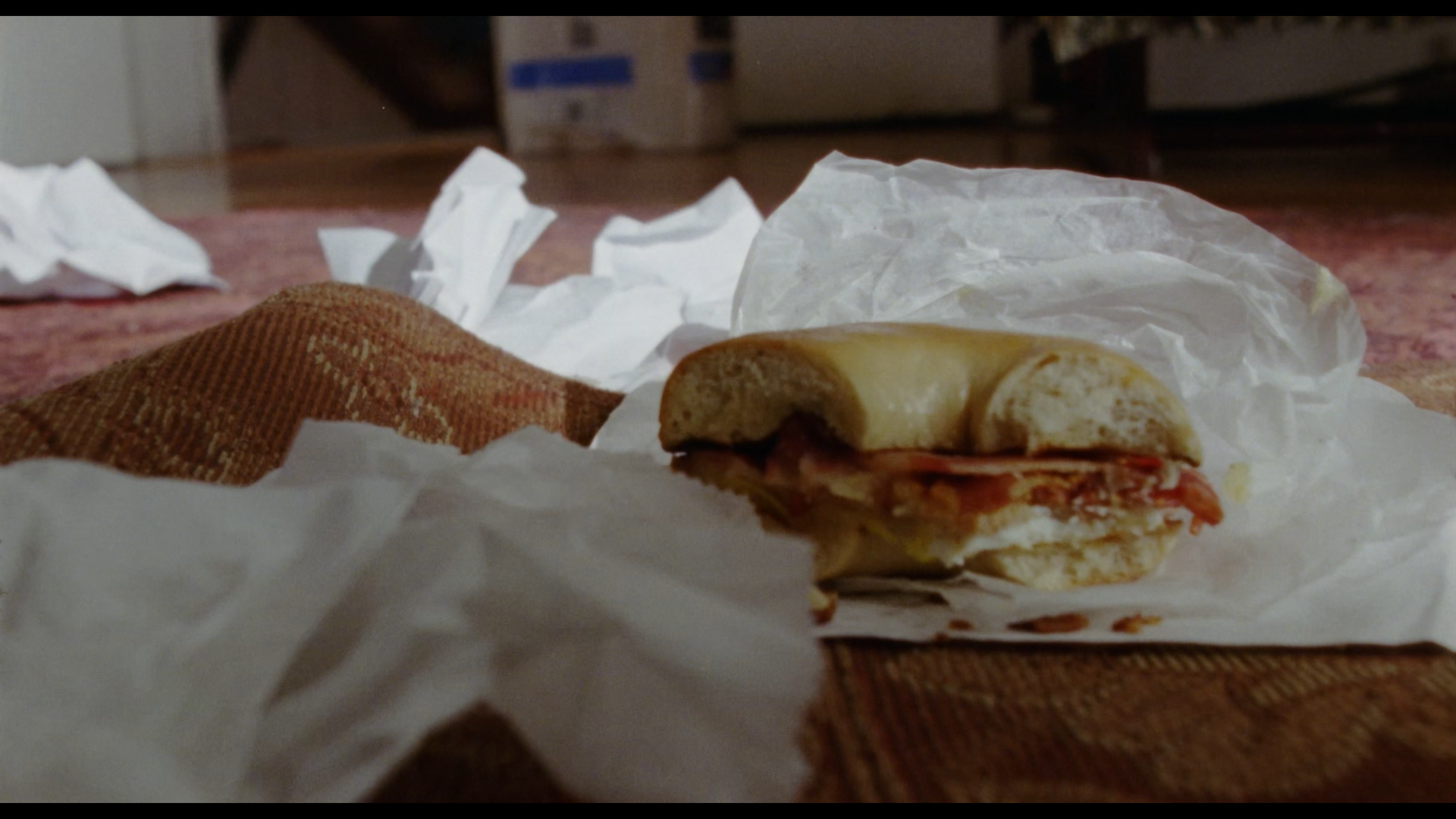 A partially eaten sandwich on crumpled white paper, with torn crumpled paper around it, on a carpeted floor in a room with visible background.