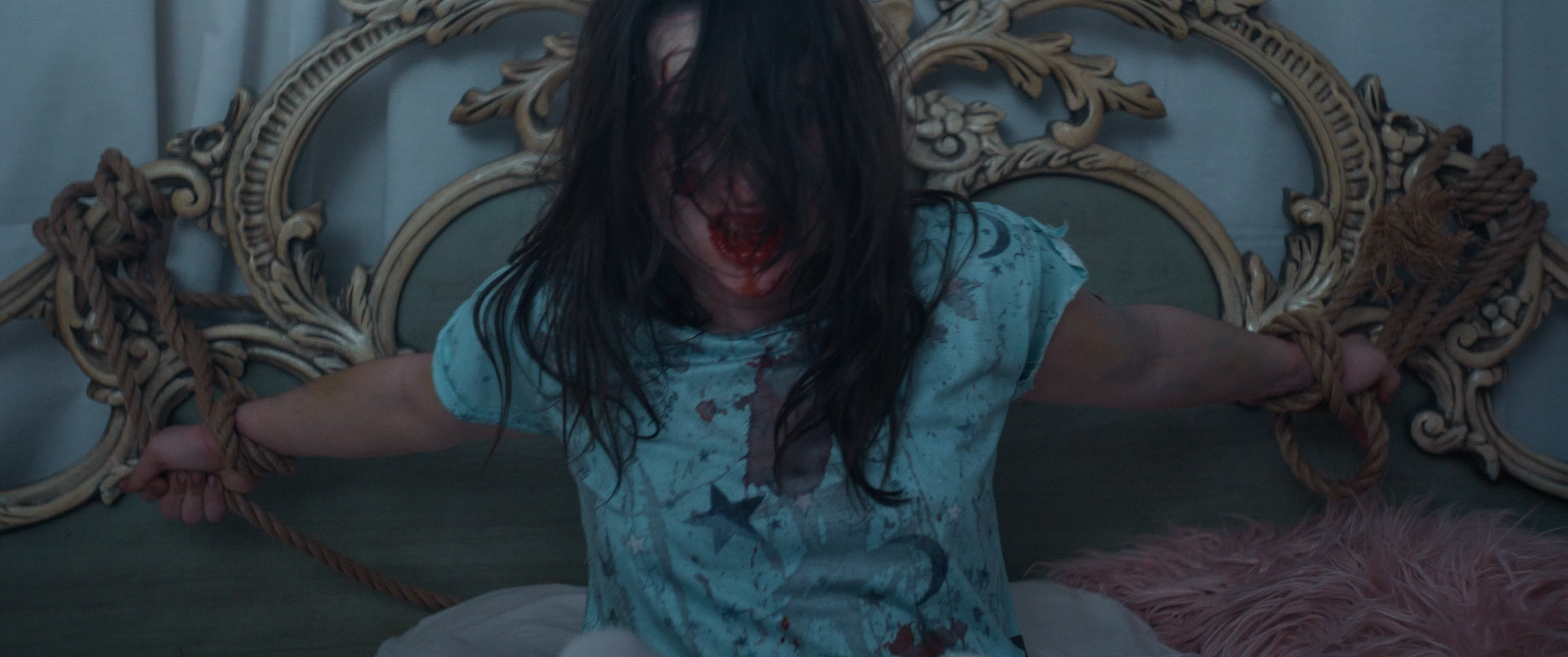 A woman with long dark hair and blood on her face and shirt, tied to a bed with thick ropes, in a dimly lit room with ornate headboard and pink furry pillow.