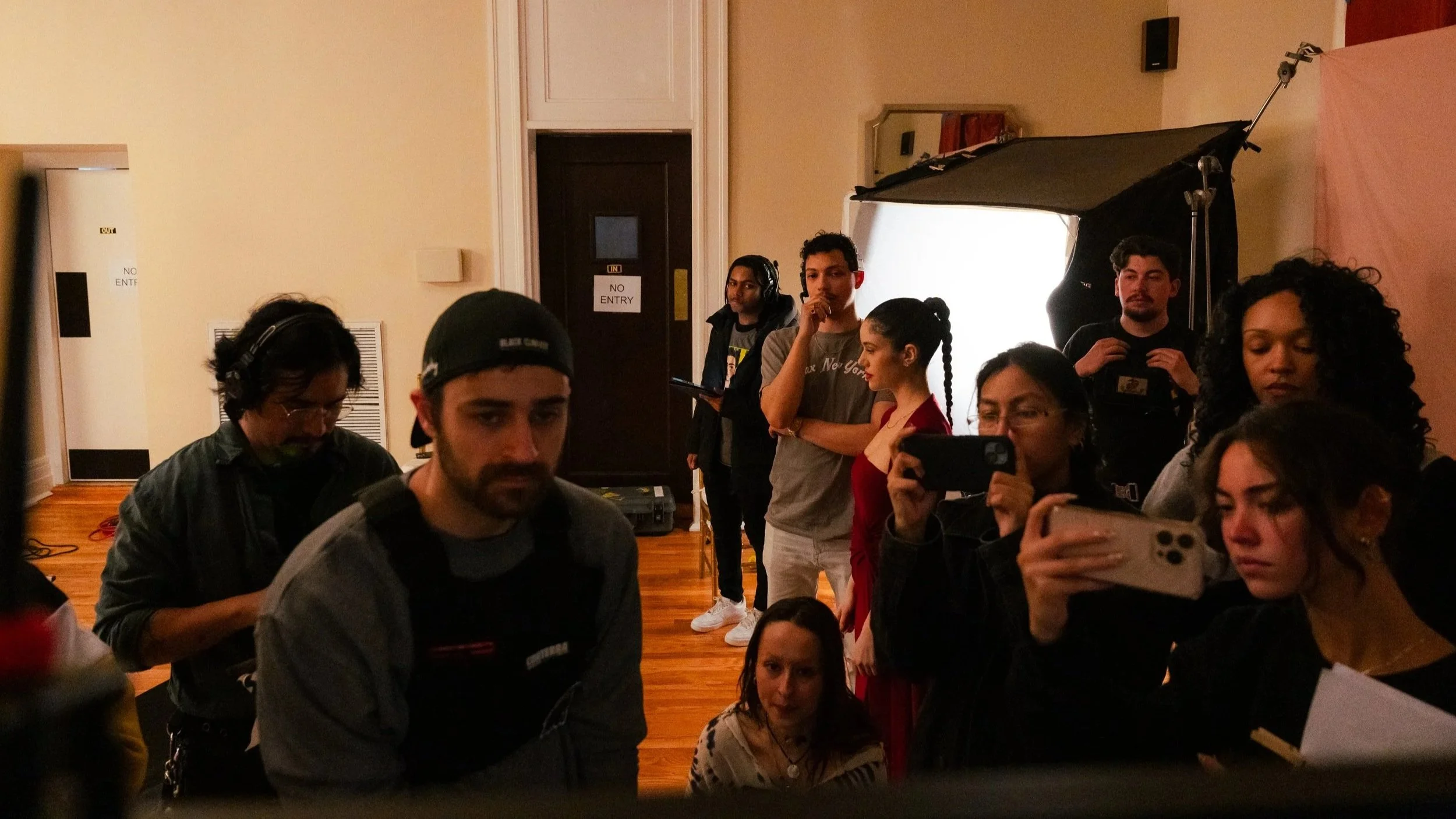 Group of people watching a filming or photoshoot in a room with wooden floors, some holding phones and taking pictures. There is studio lighting equipment in the background and