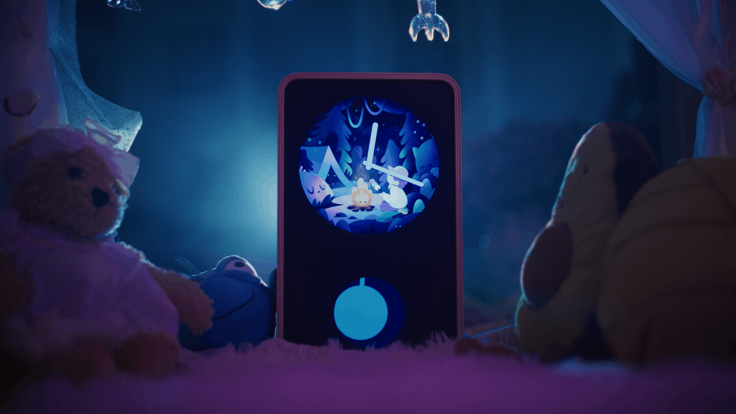 A digital clock displayed in a children's bedroom with stuffed animals, including a teddy bear and a duck, around it. The clock shows a cartoon camping scene with a campfire, trees, and animals, with a night sky background.