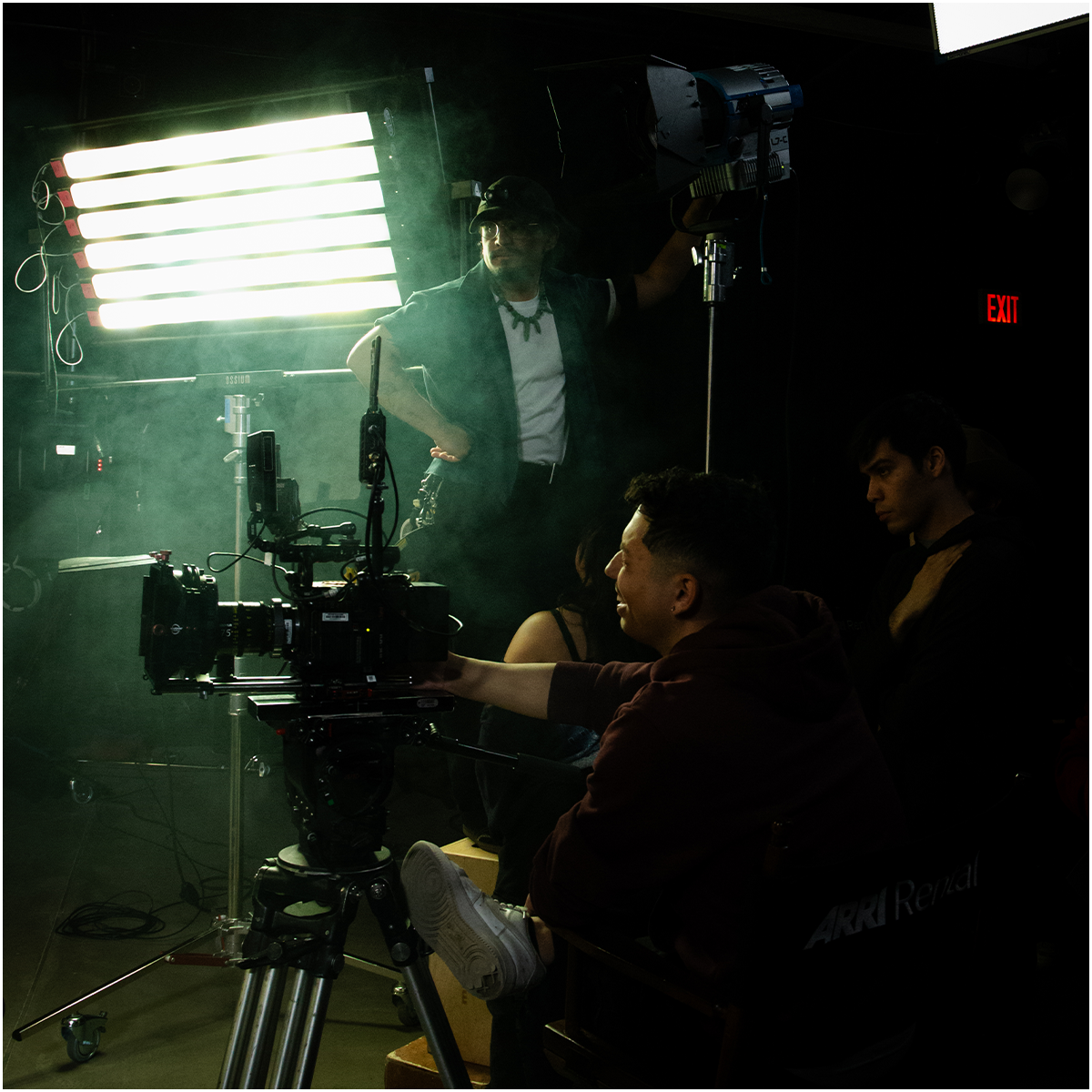Production crew on set with camera and lighting equipment, in a dark studio with green smoke or haze, illuminated by a rectangular LED light panel, behind the scenes of a filming or photography session.