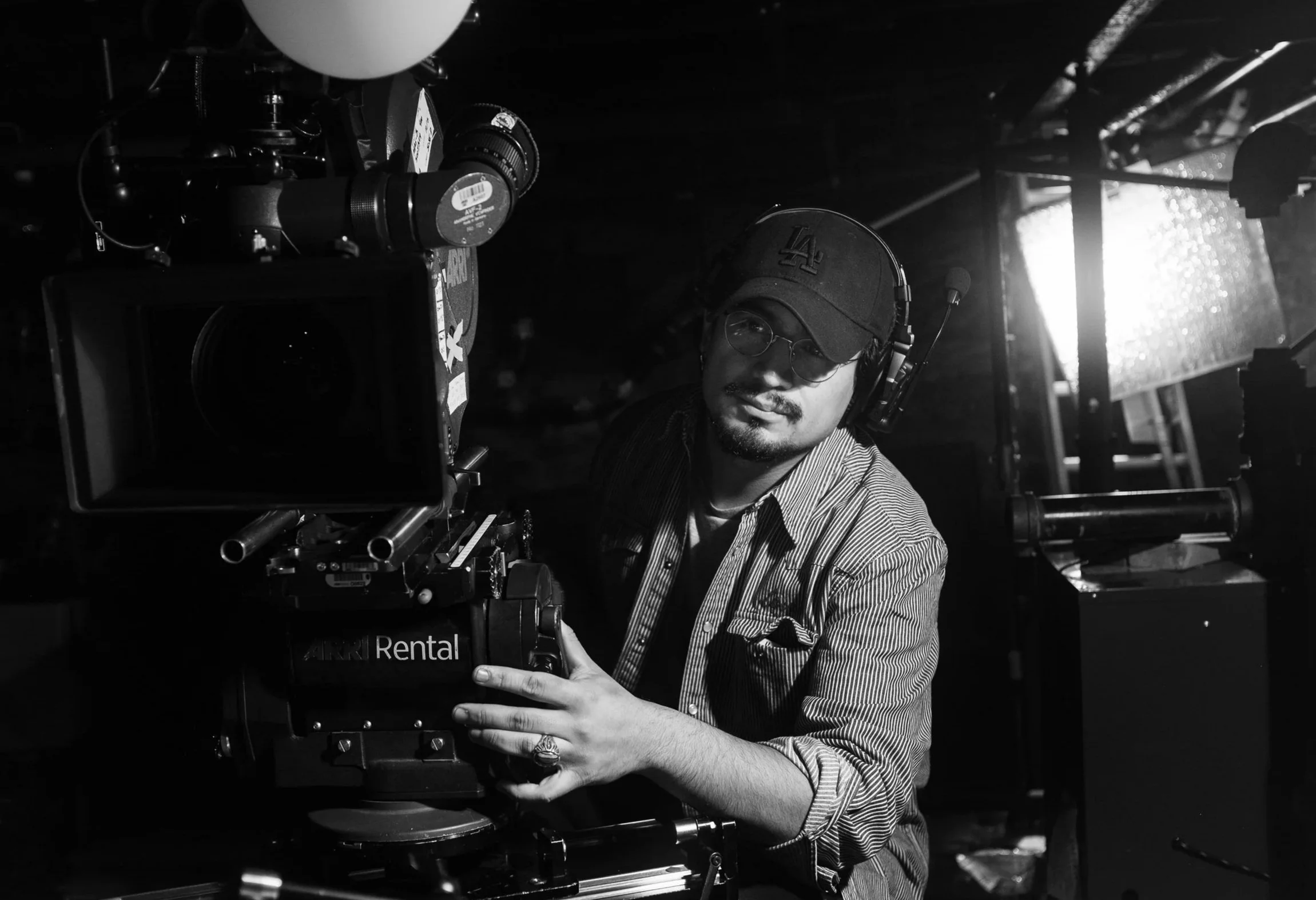 A man wearing glasses, a cap, and headphones operating a professional movie camera on a film set, with a bright light in the background.