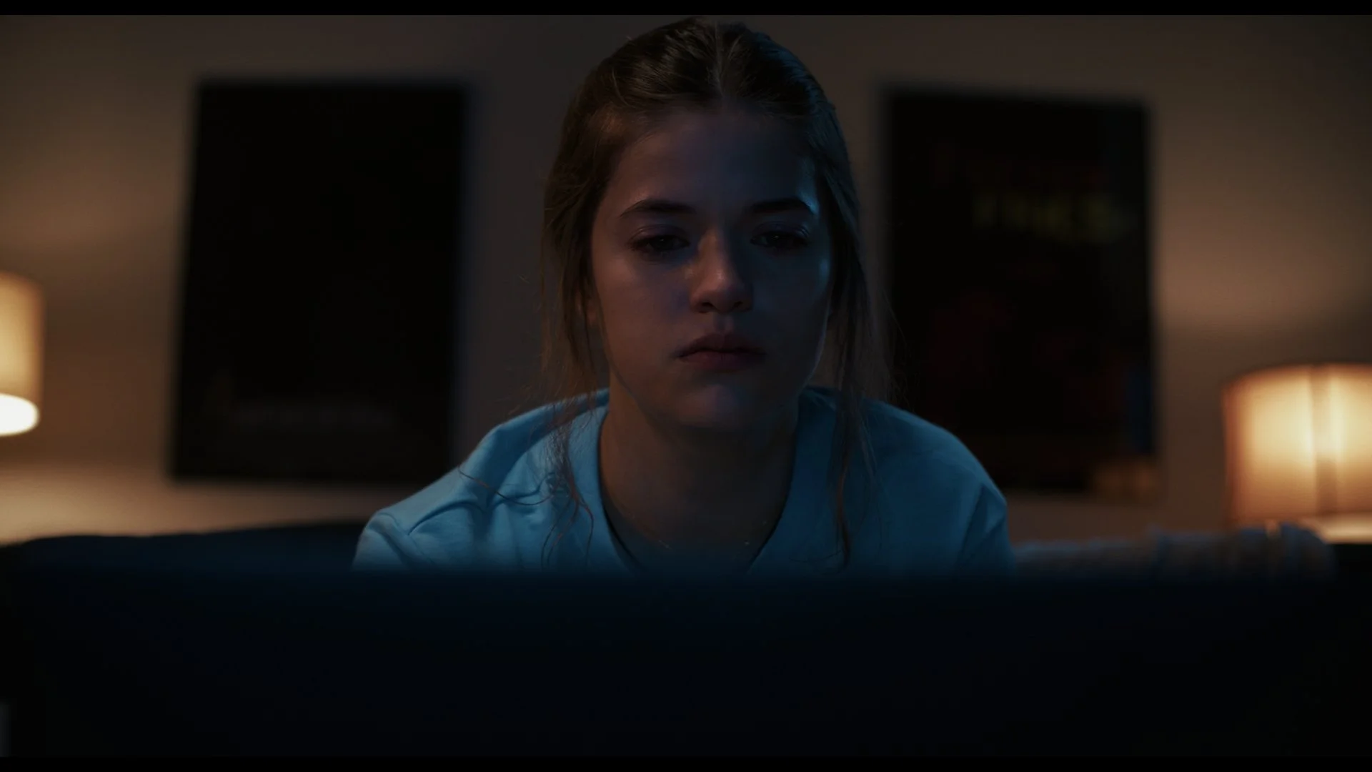 A young woman with wet hair is sitting in a dimly lit room, staring intently at a screen in front of her, with two lamps providing warm light in the background.