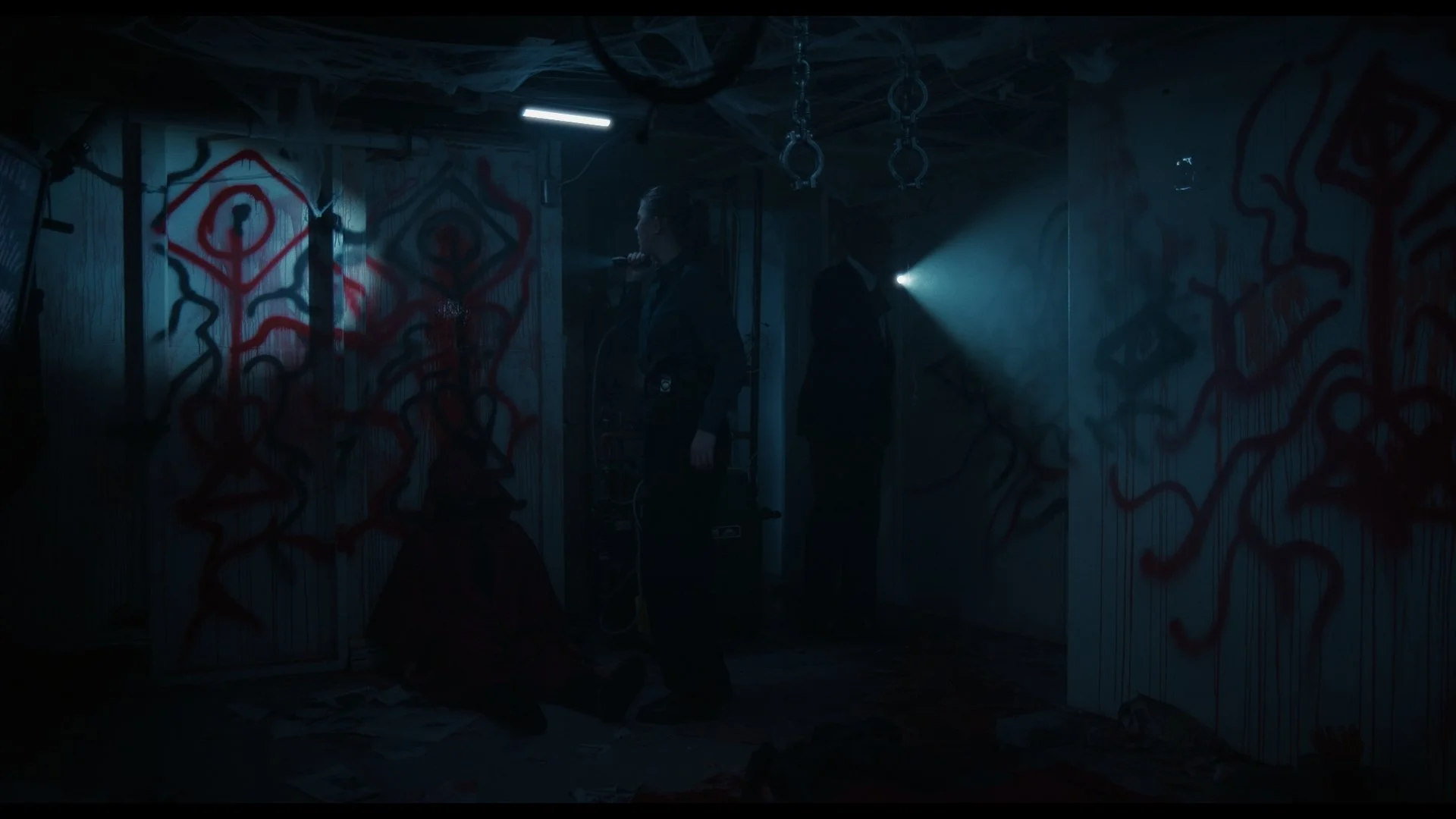 Dark, eerie basement with graffiti-covered walls, featuring a person holding a flashlight, another person in black, and various hanging gymnastic rings.