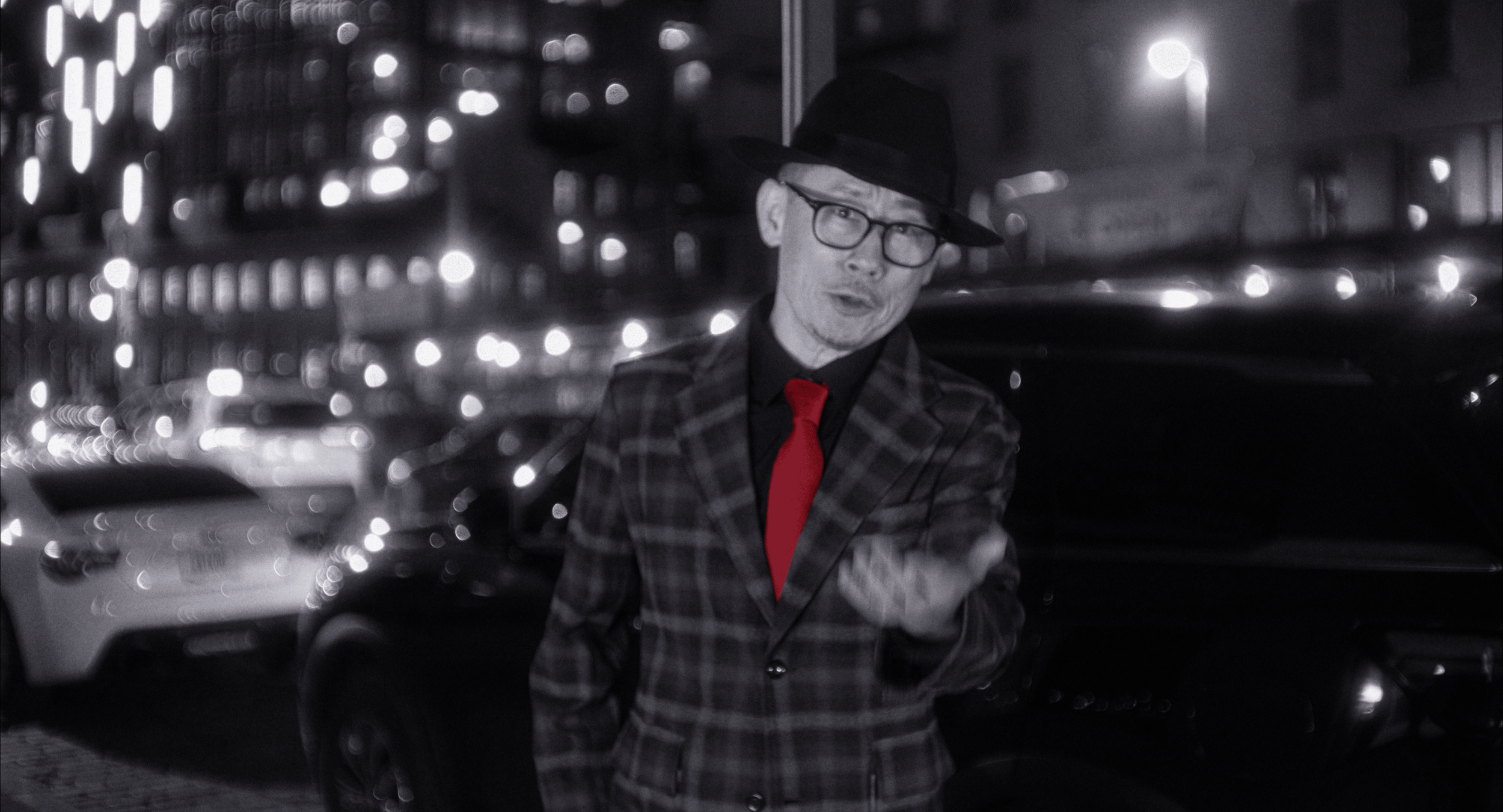 A man wearing glasses, a black hat, a plaid suit, and a red tie stands in a city street at night, with blurred city lights and parked cars in the background.