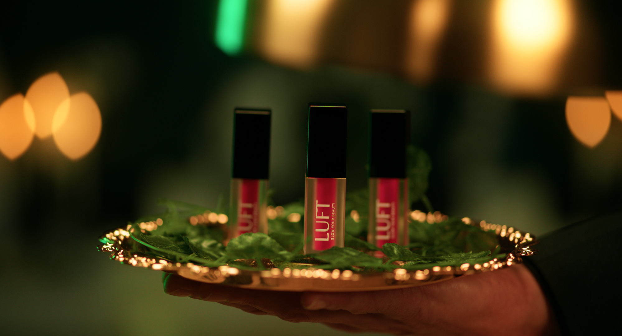 Three pink and black lip gloss tubes labeled 'LUVF,' placed on a gold tray with green decorative leaves, with blurred warm lighting in the background.