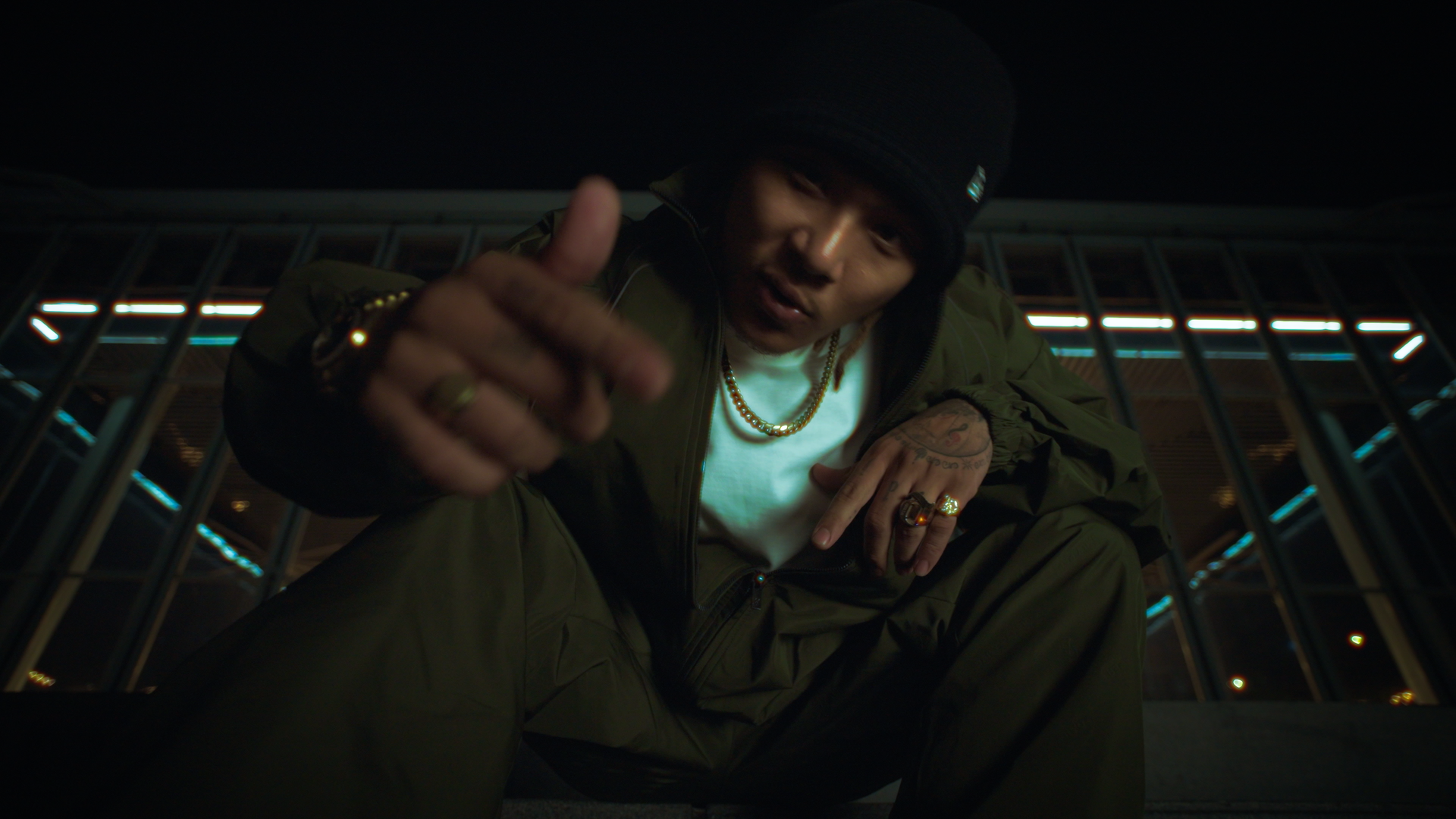 A man wearing a black beanie, green jacket, and gold jewelry is crouching on the ground at night, pointing toward the camera with his right hand, under a metallic structure with blue and white lights.