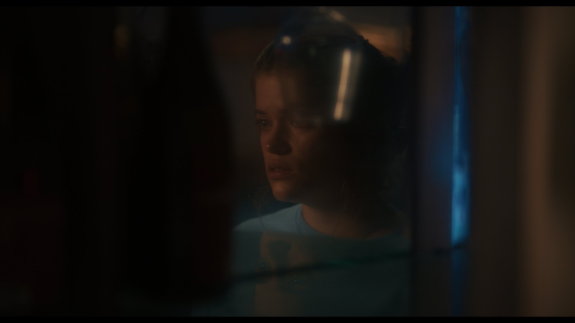 A woman seen through a partially opened fridge door, with the fridge light illuminating her face in a dark setting.