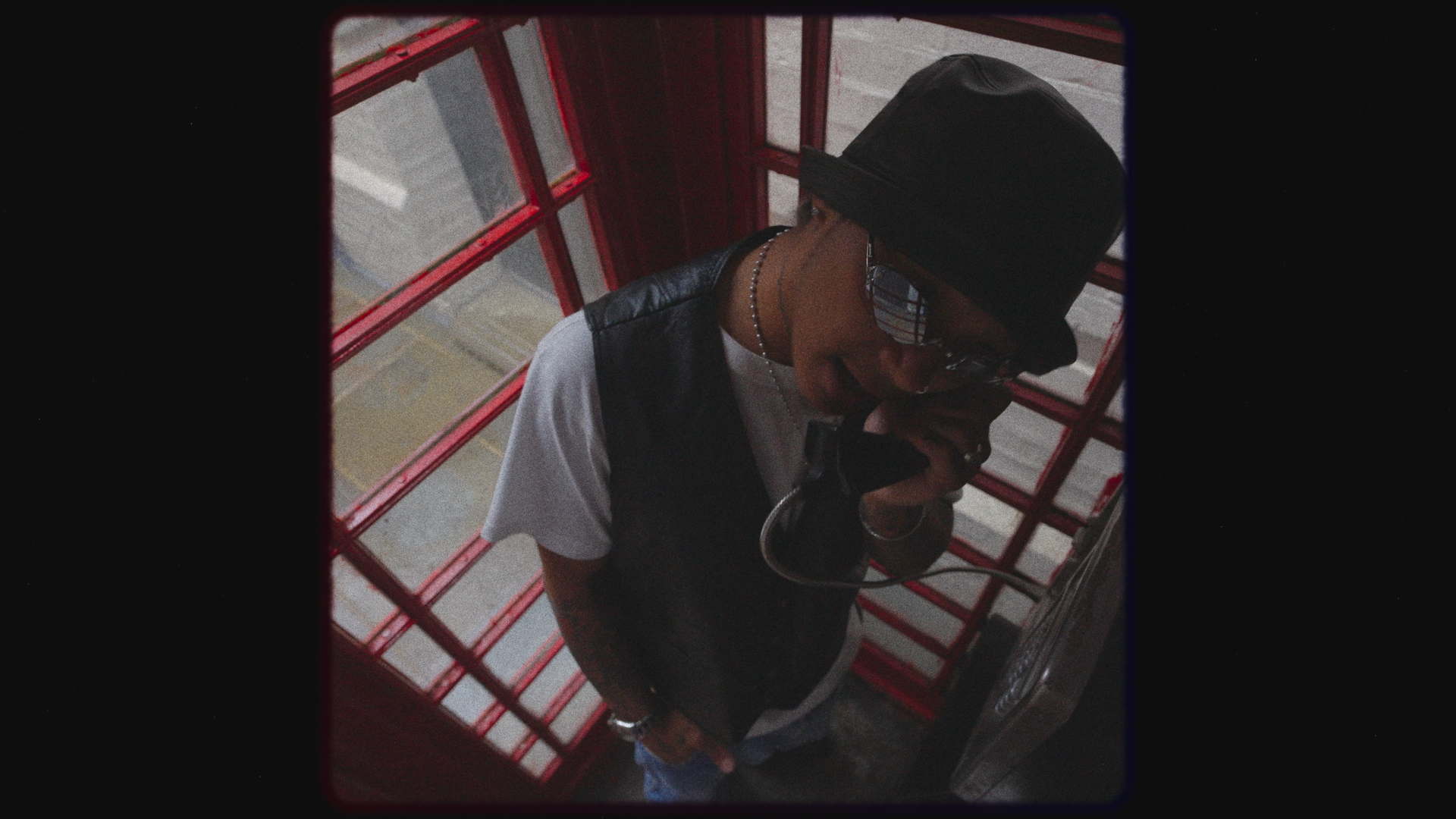 A young person taking a selfie in a red phone booth, wearing a black bucket hat, sunglasses, a black vest over a white t-shirt, with jewelry, holding the phone close to their face.