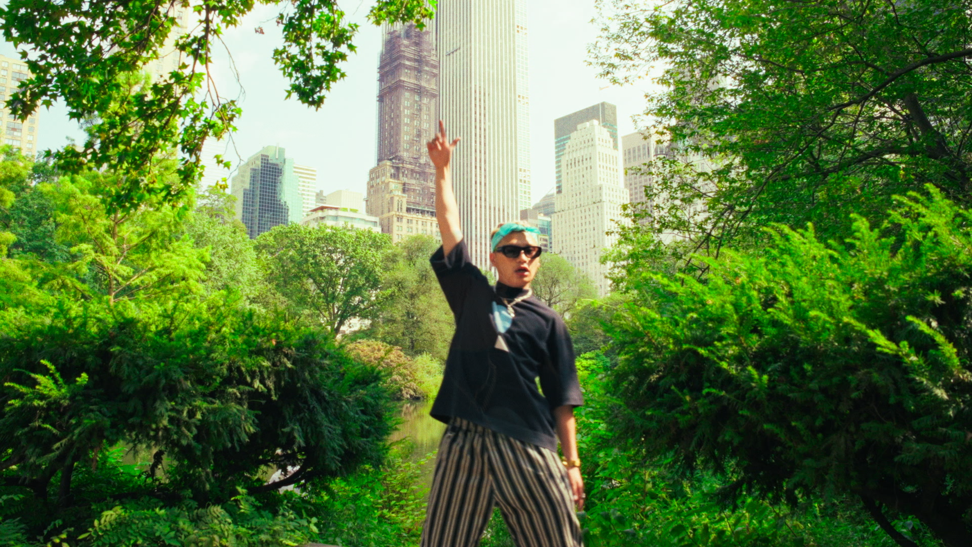 Person wearing sunglasses and striped pants posing outdoors in a park surrounded by green trees with tall city buildings in the background.